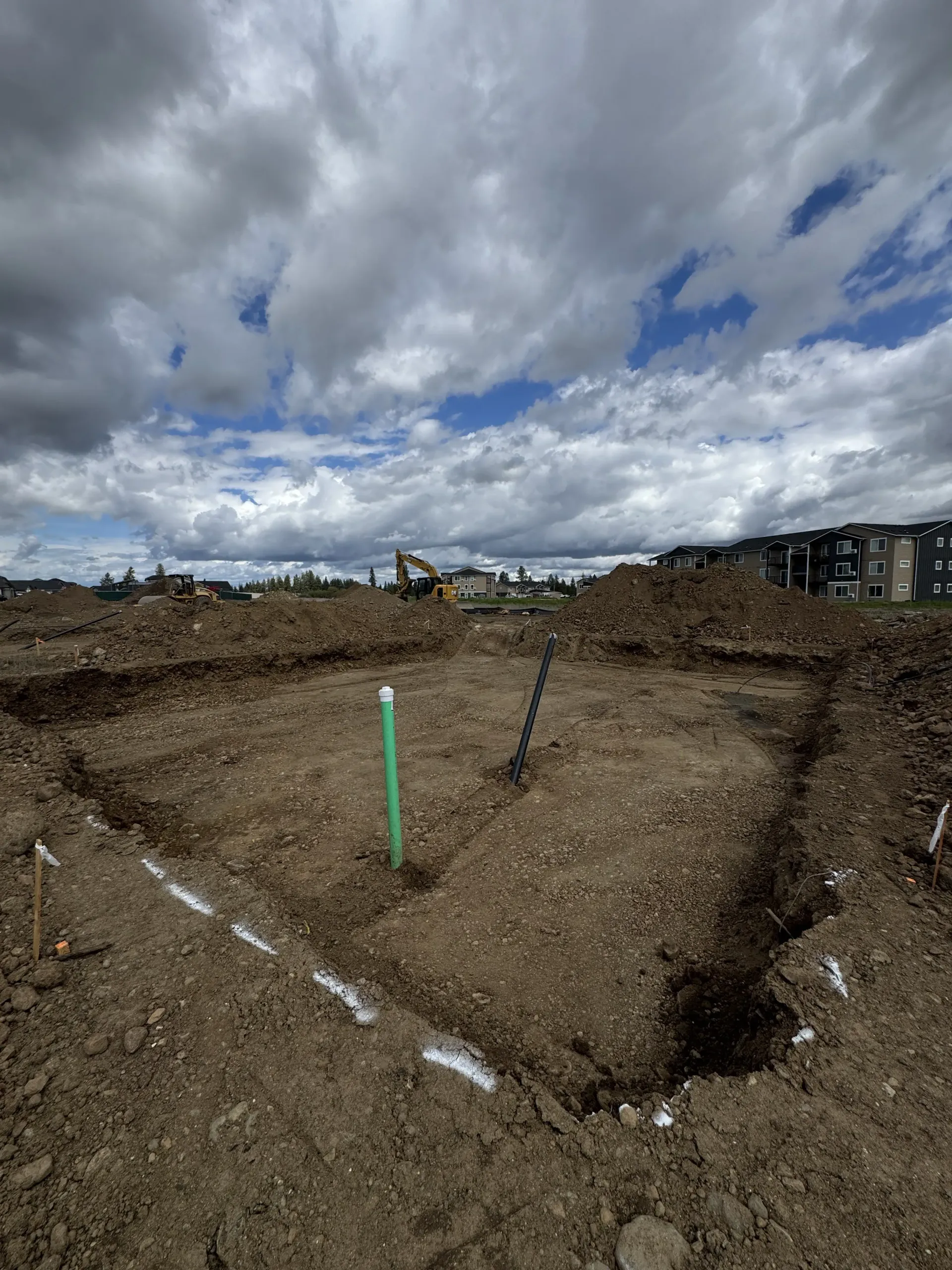 A rectangular excavation in a dirt field with a green pipe sticking out, under a cloudy sky.