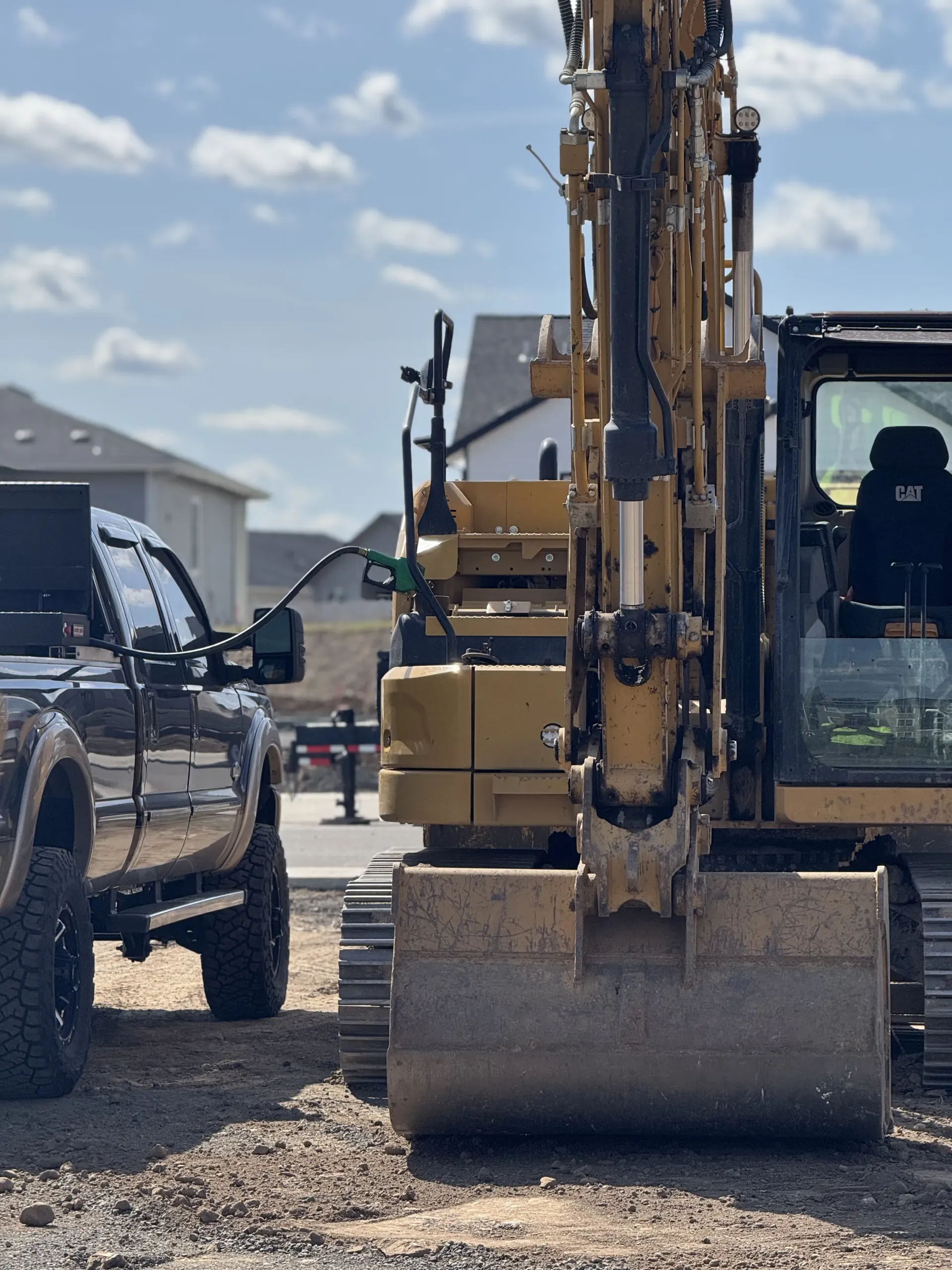 A black pickup truck parked next to a large yellow excavator on a construction site under a partly cloudy sky.