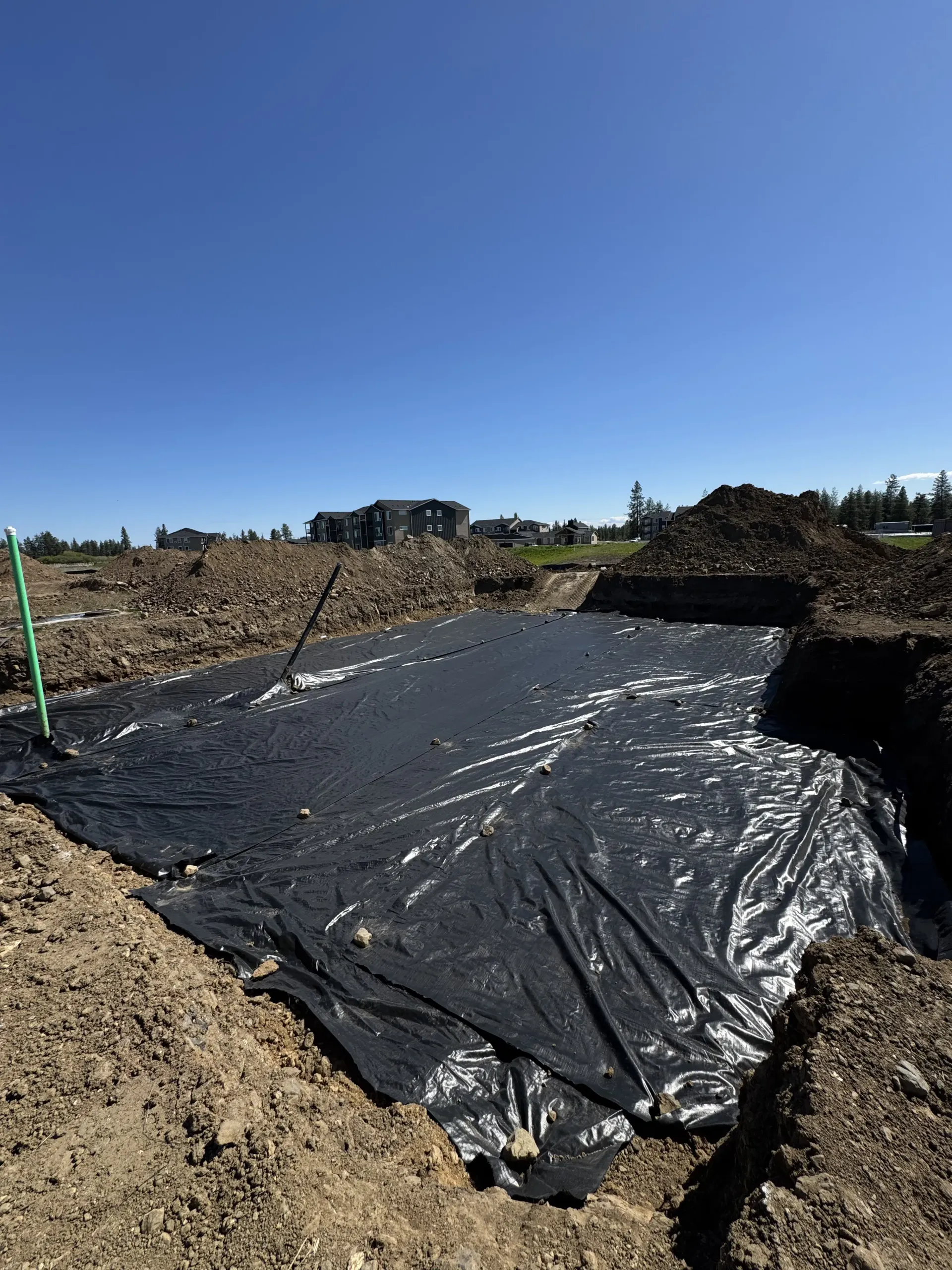A large black plastic liner covers the ground in a rectangular excavation pit at a construction site under a blue sky.