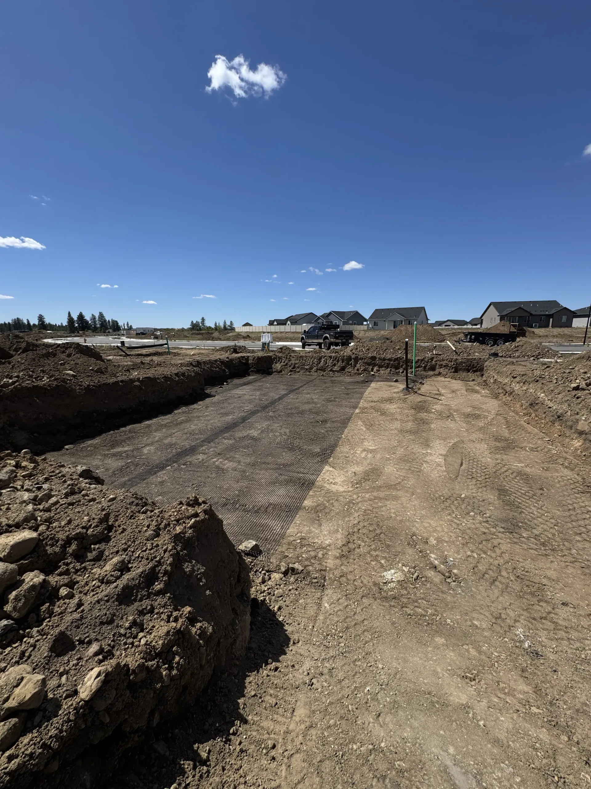 A construction site featuring an excavated trench lined with a black geogrid mesh under a clear blue sky.
