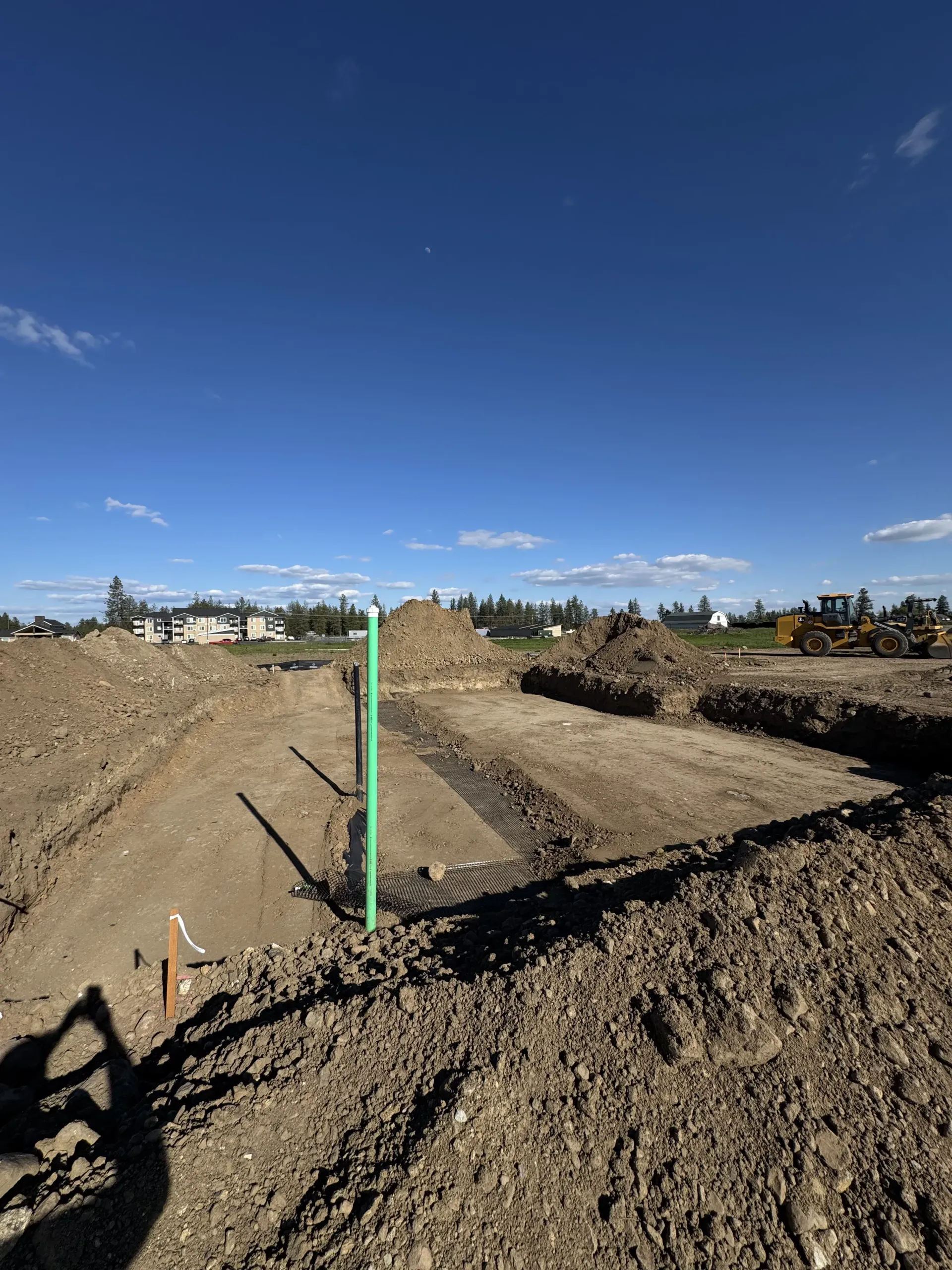 A construction site with excavated trenches, piles of dirt, and a green survey marker under a bright blue sky.