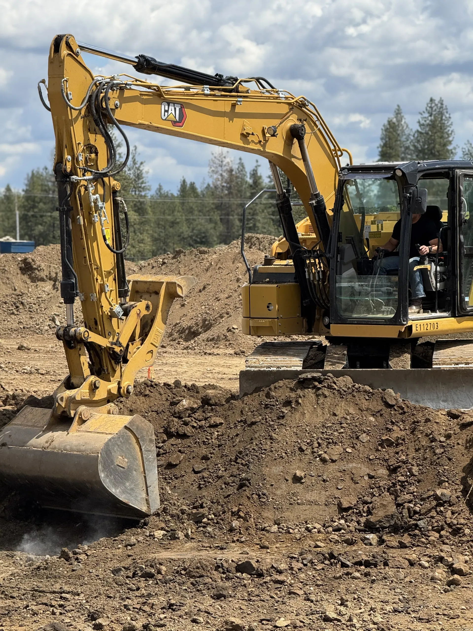 A yellow Caterpillar excavator digging in a dirt lot under a cloudy sky with an operator inside the cab.