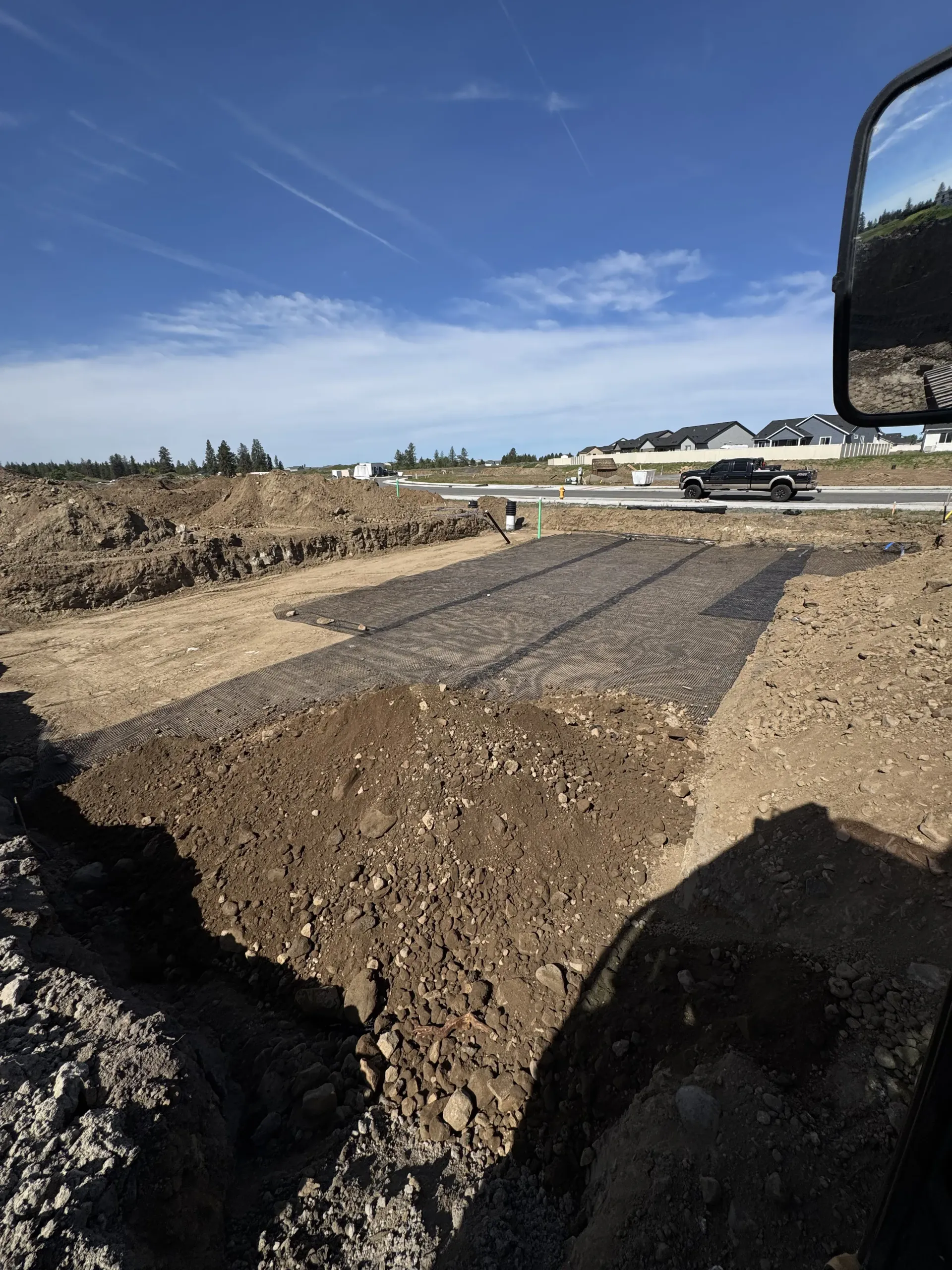 A construction site showing a patch of black geogrid material laid out on graded dirt under a clear blue sky.