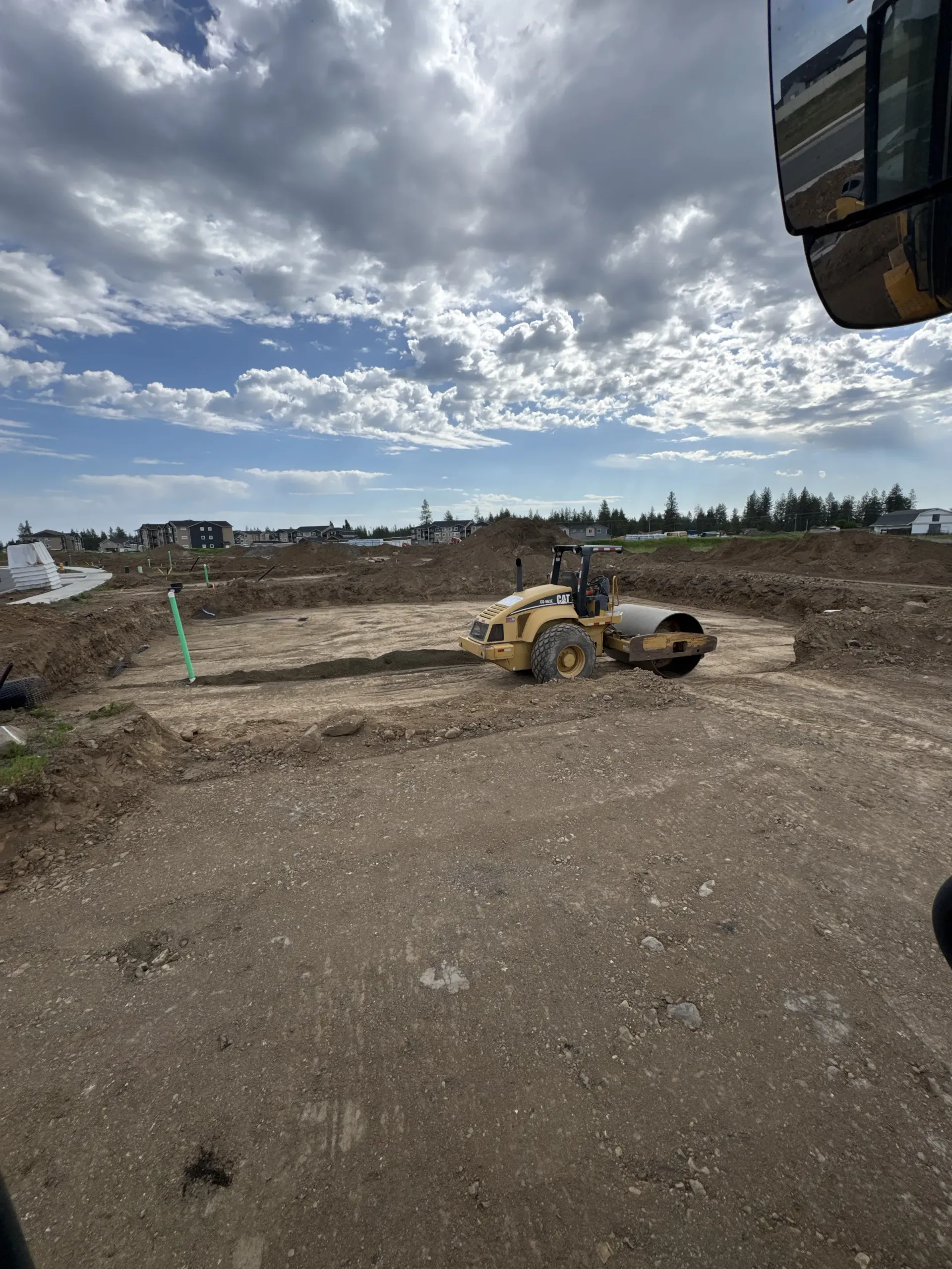 A yellow soil compactor operates on a dirt construction site under a bright, cloudy sky.