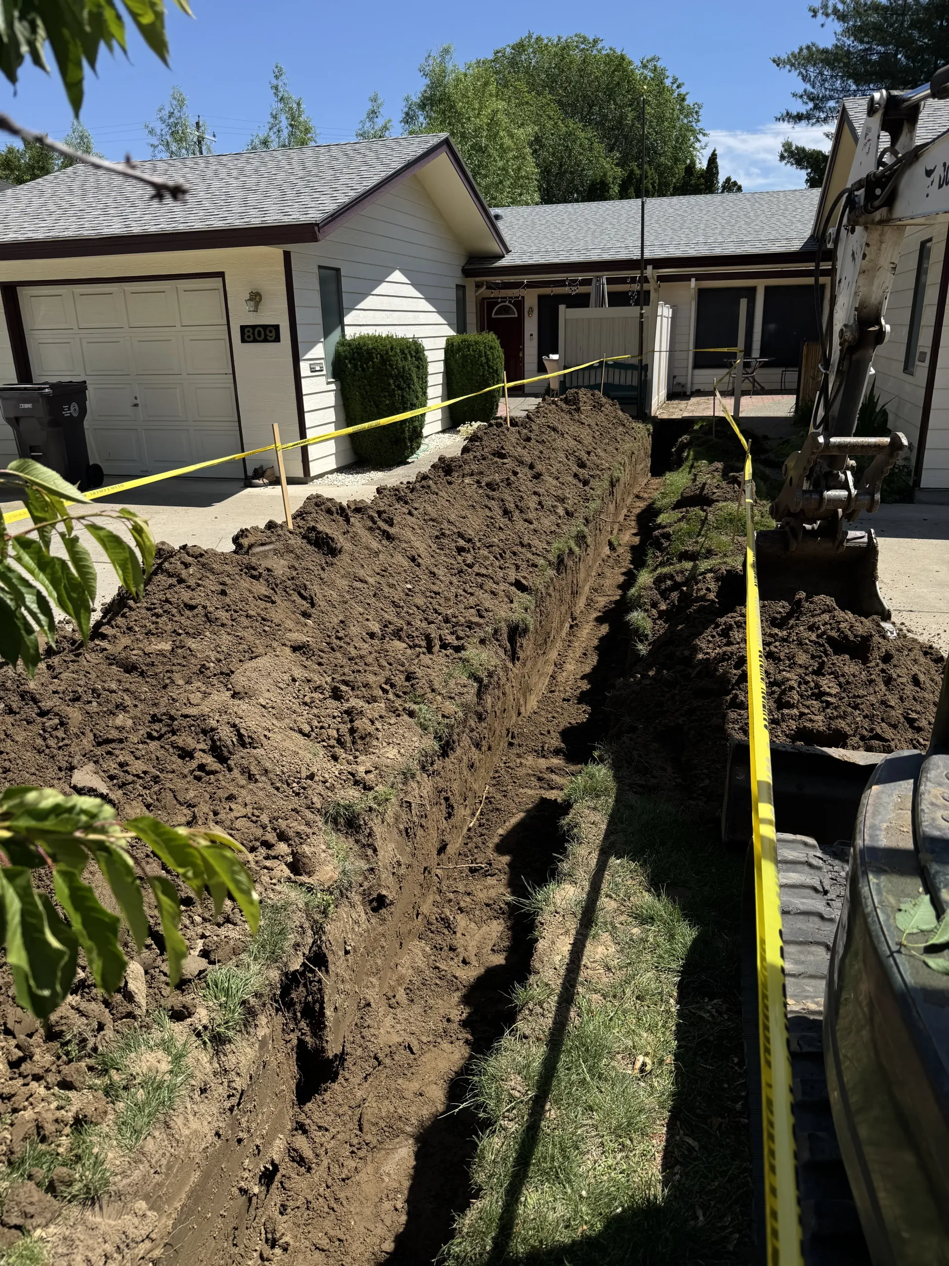 A deep trench dug for construction in a residential yard, bordered by yellow caution tape with a house in the background.