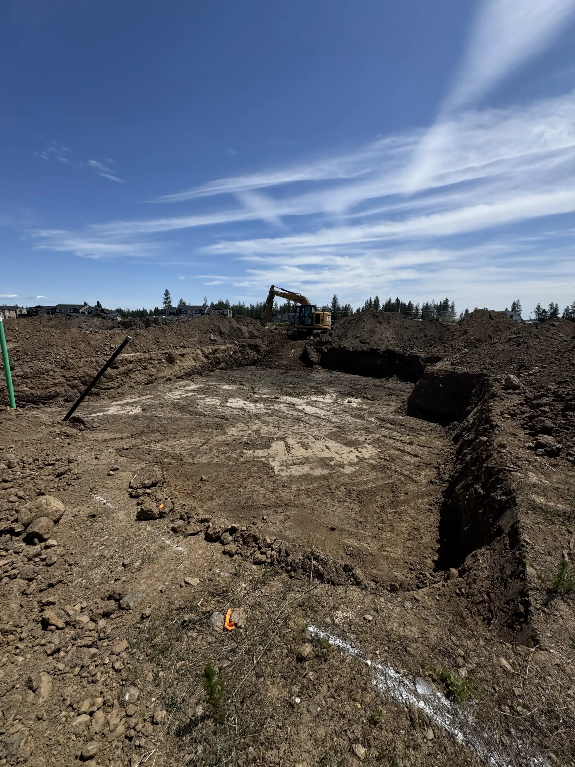 An excavator sits in a large, dirt-filled construction excavation site under a bright blue sky.