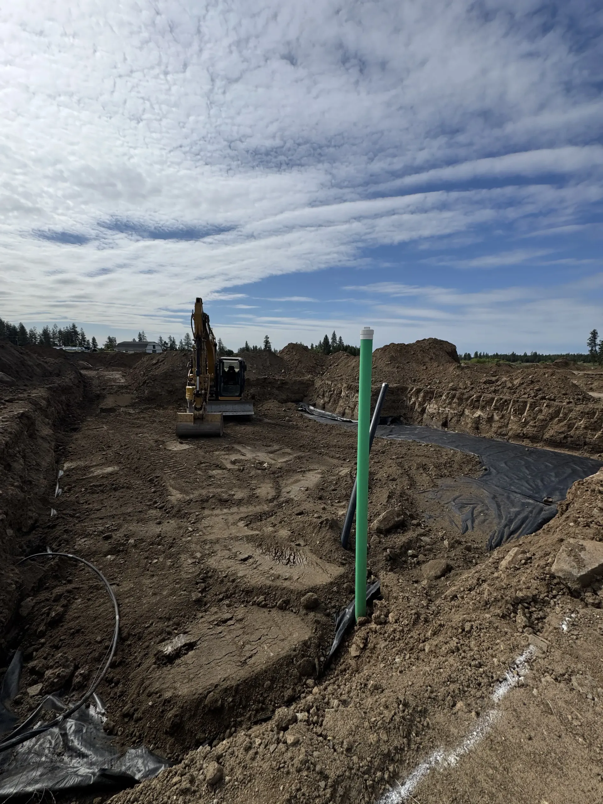 Construction site with an excavator, a vertical green pipe, and black ground cover in a dirt trench under a cloudy sky.