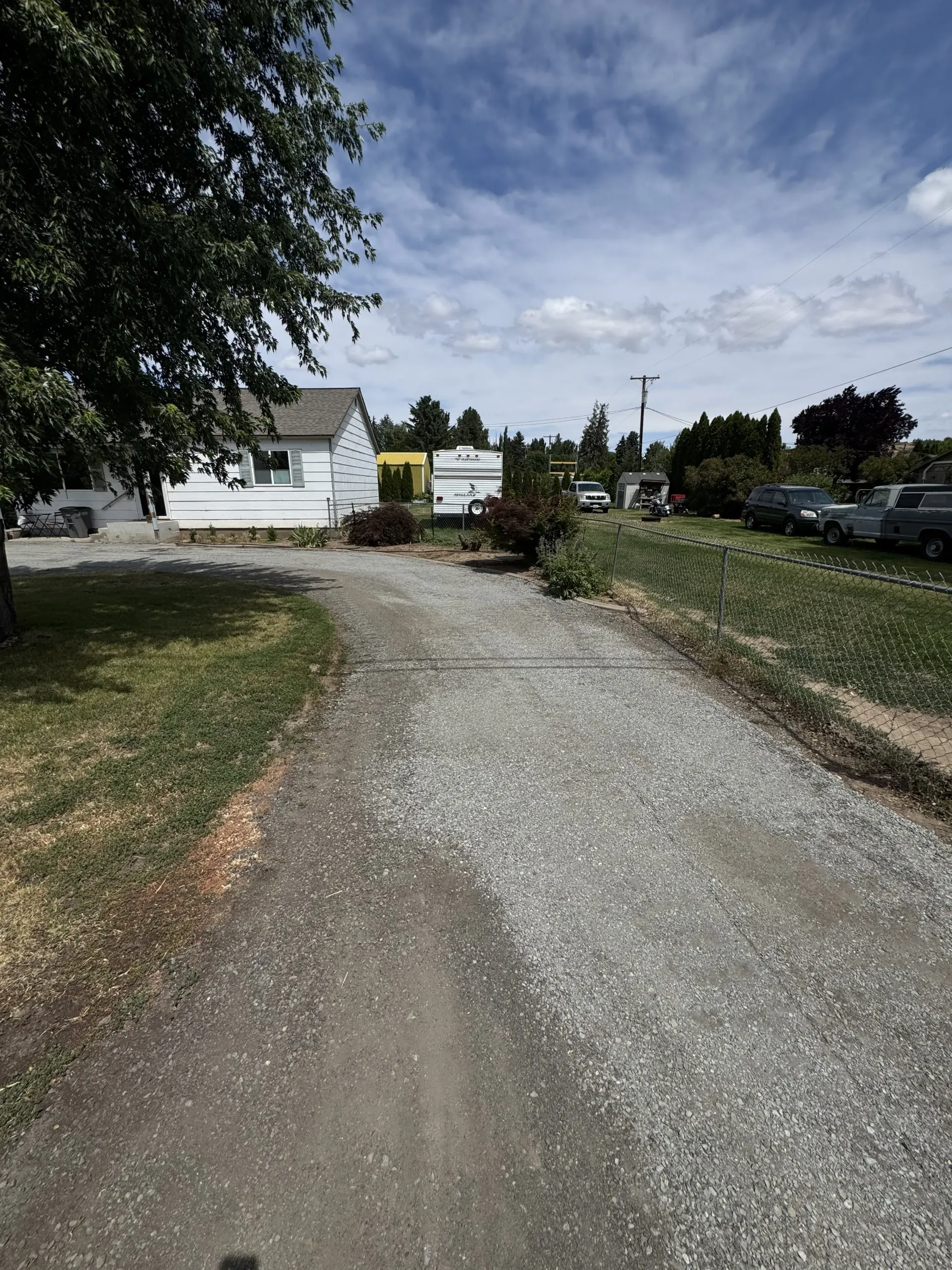 A gravel driveway leads to a white house under a blue, cloudy sky, with several parked vehicles visible in the distance.