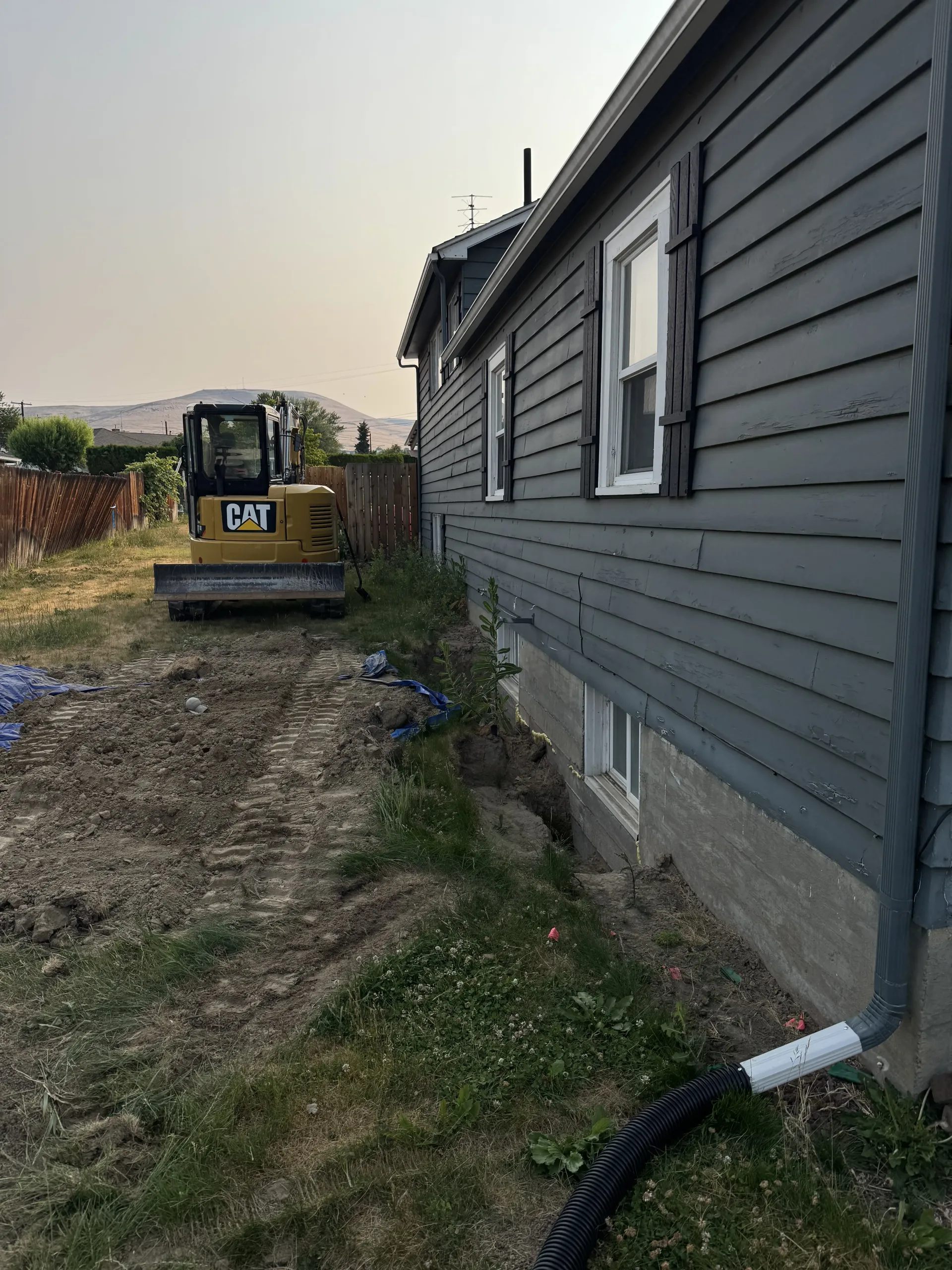 A yellow excavator parked near a house with gray siding and an exposed foundation trench in a grassy backyard.