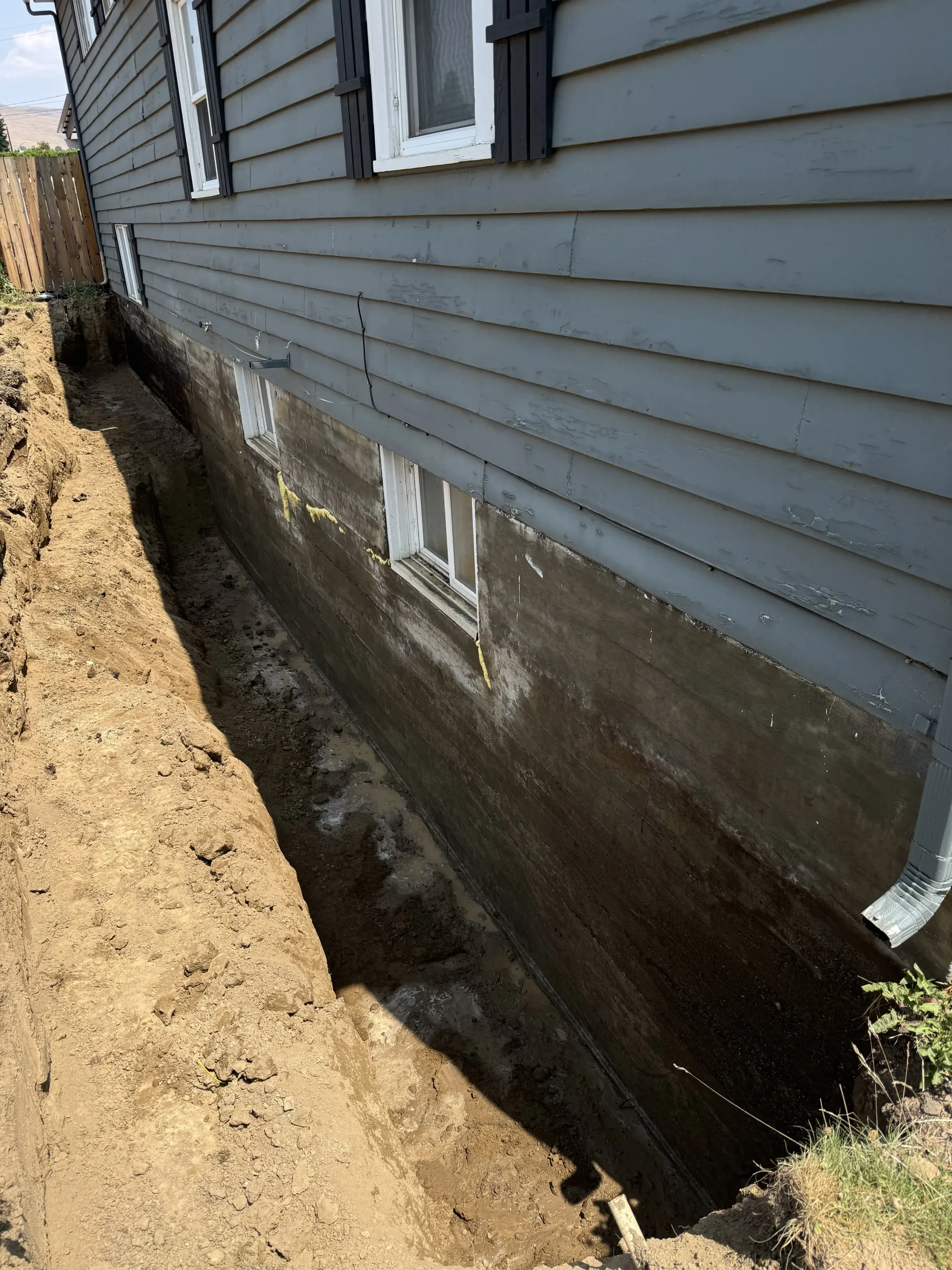 A narrow trench dug along the gray-sided exterior foundation of a house, exposing a freshly waterproofed concrete wall.