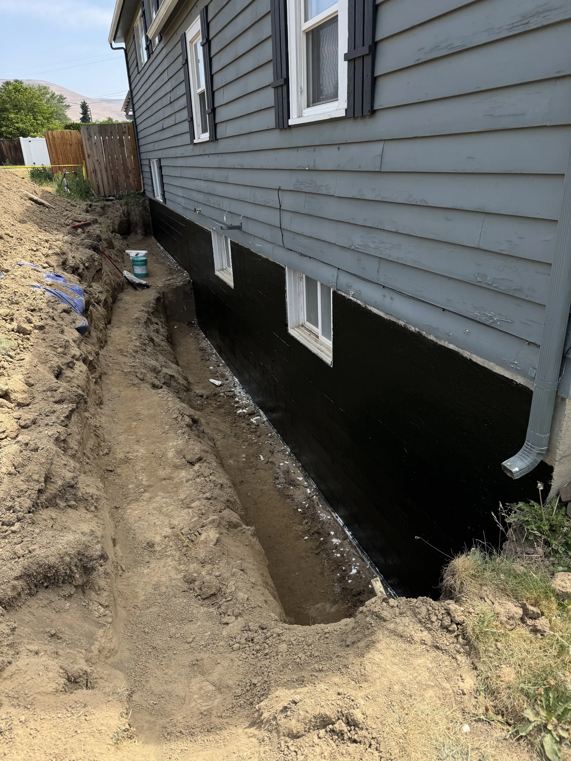 A freshly excavated trench alongside the black, waterproofed foundation wall of a grey-sided house.