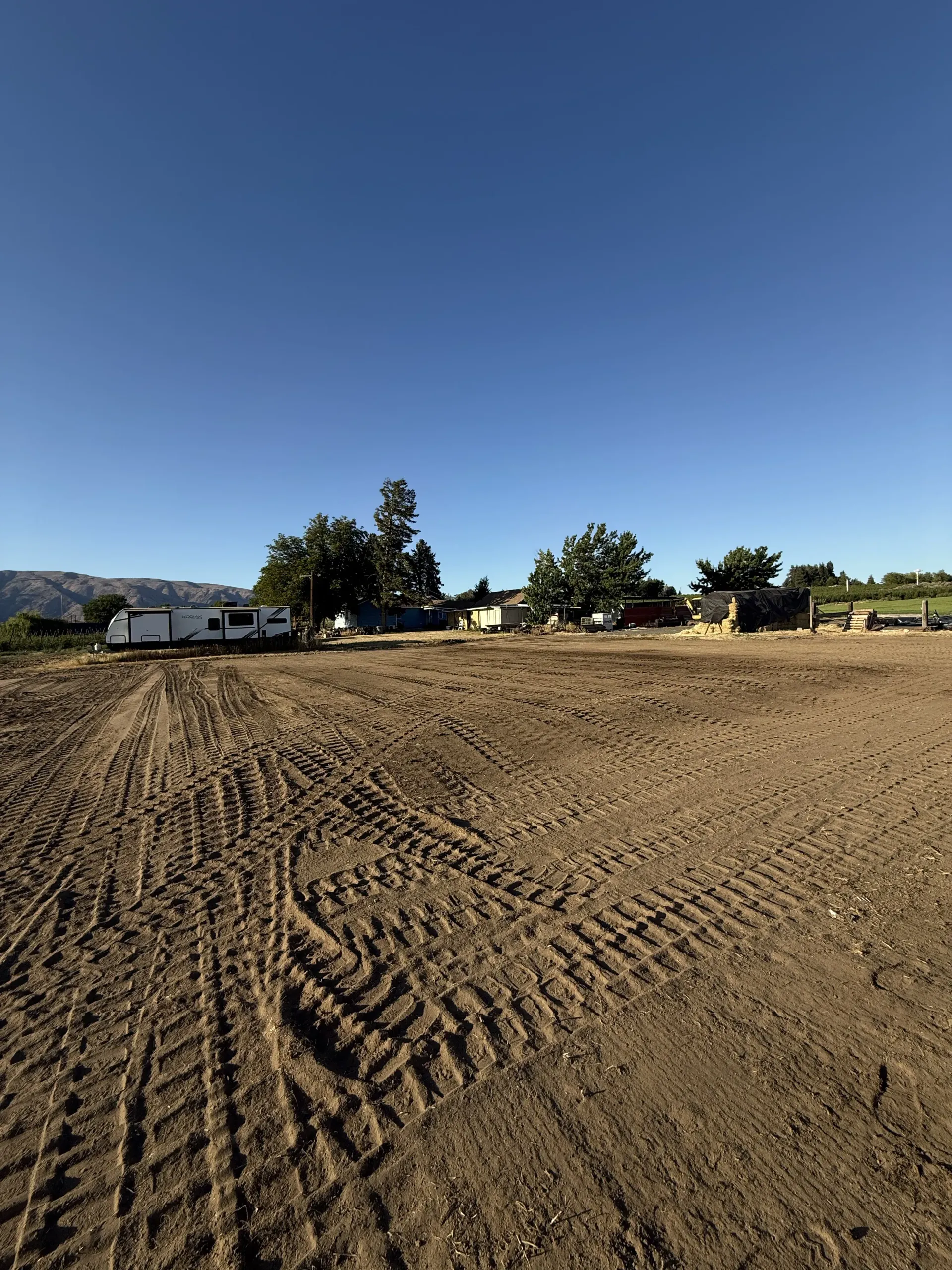 A wide, dirt-covered field with tire tracks under a clear blue sky, featuring a distant trailer and trees.