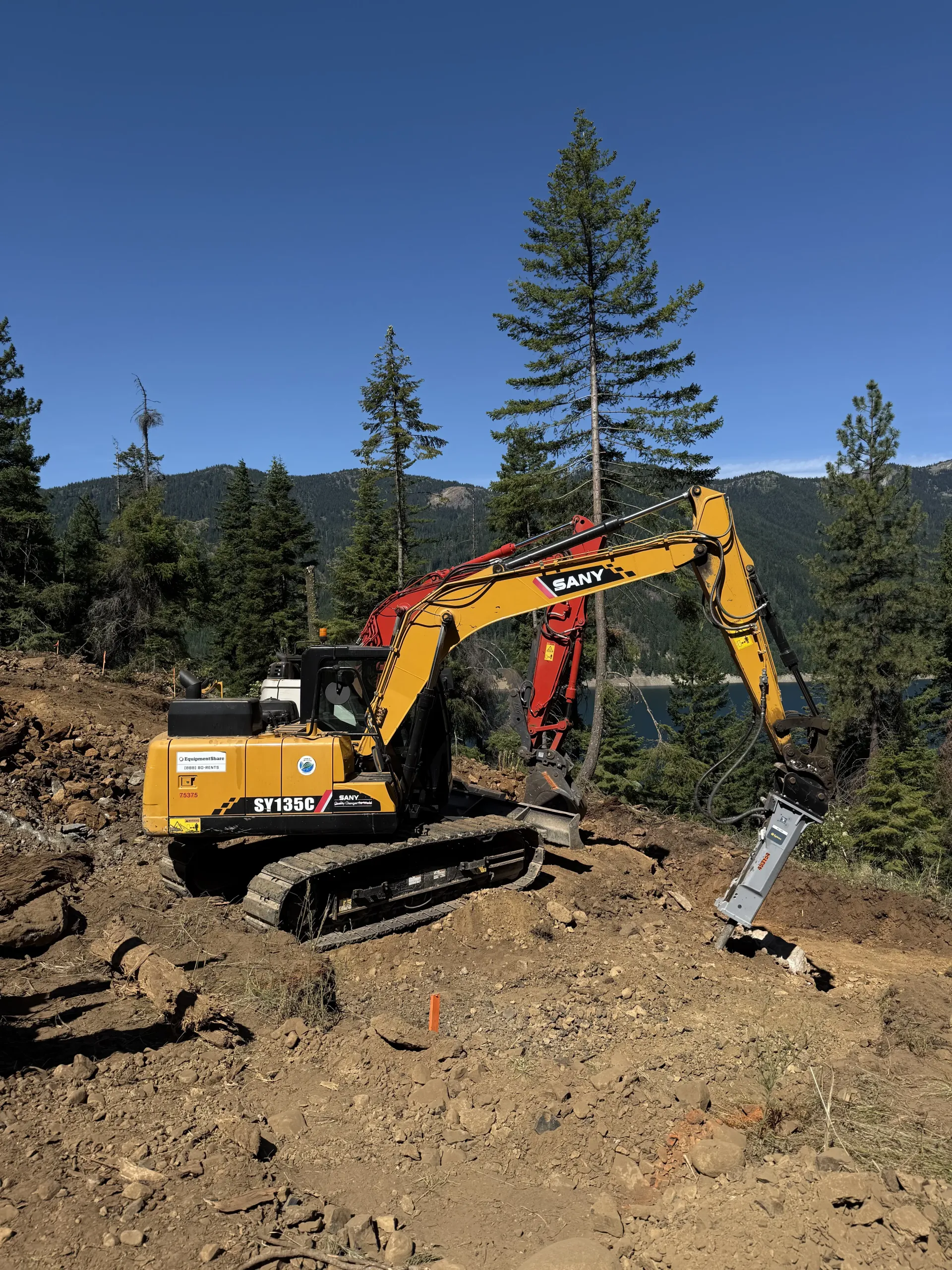 A yellow John Deere excavator with a hydraulic hammer attachment sits on a rocky slope in a forested, mountainous area.