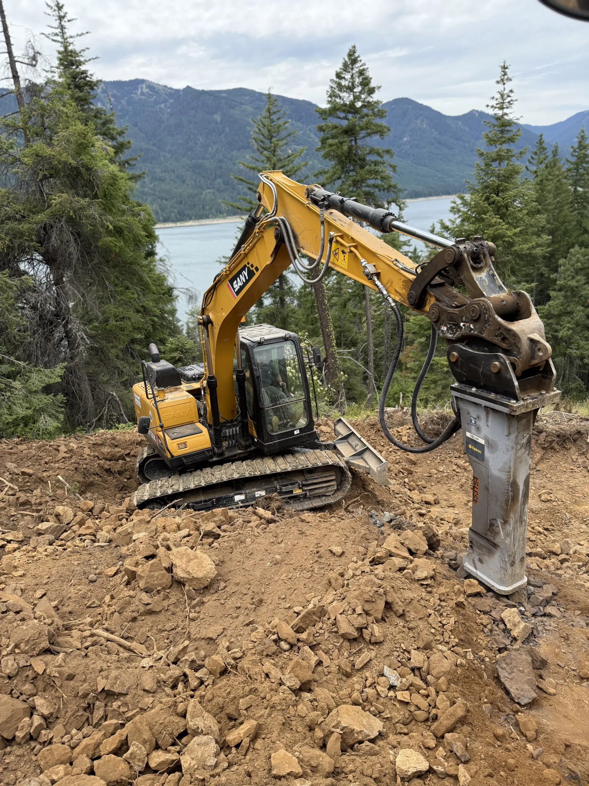 A yellow excavator with a hydraulic breaker attachment sits on a rocky hillside overlooking a lake and mountains.