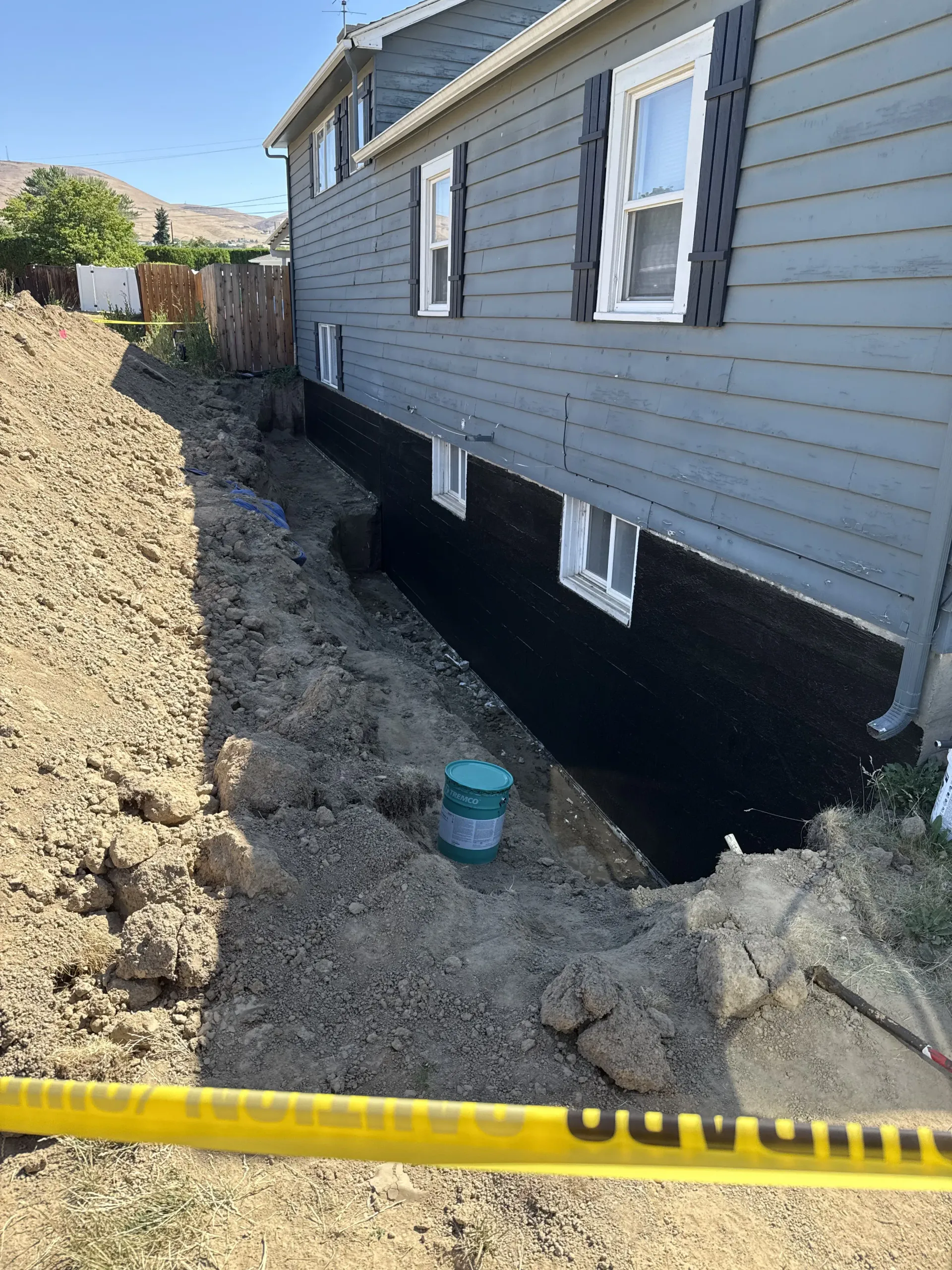 A house exterior with a foundation wall coated in black waterproofing, adjacent to a dirt trench with a green pipe cap.