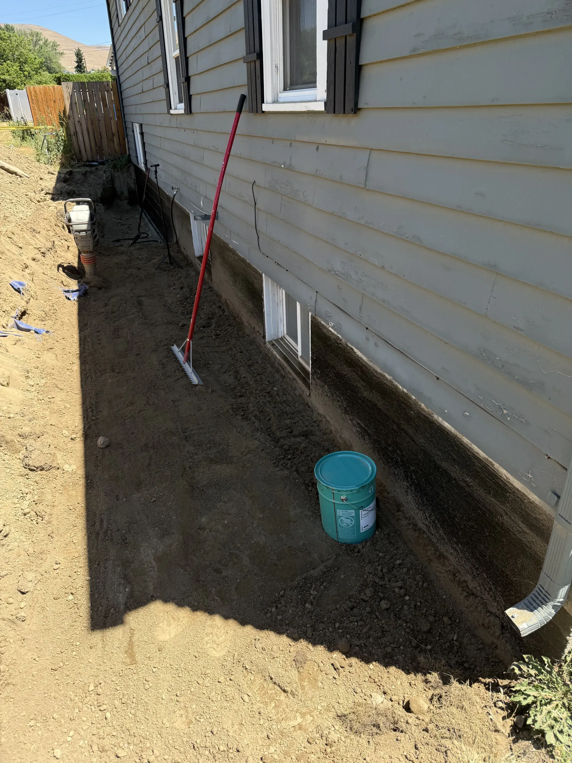 A house exterior with gray siding features a trench dug along the foundation, coated in black waterproofing sealant.