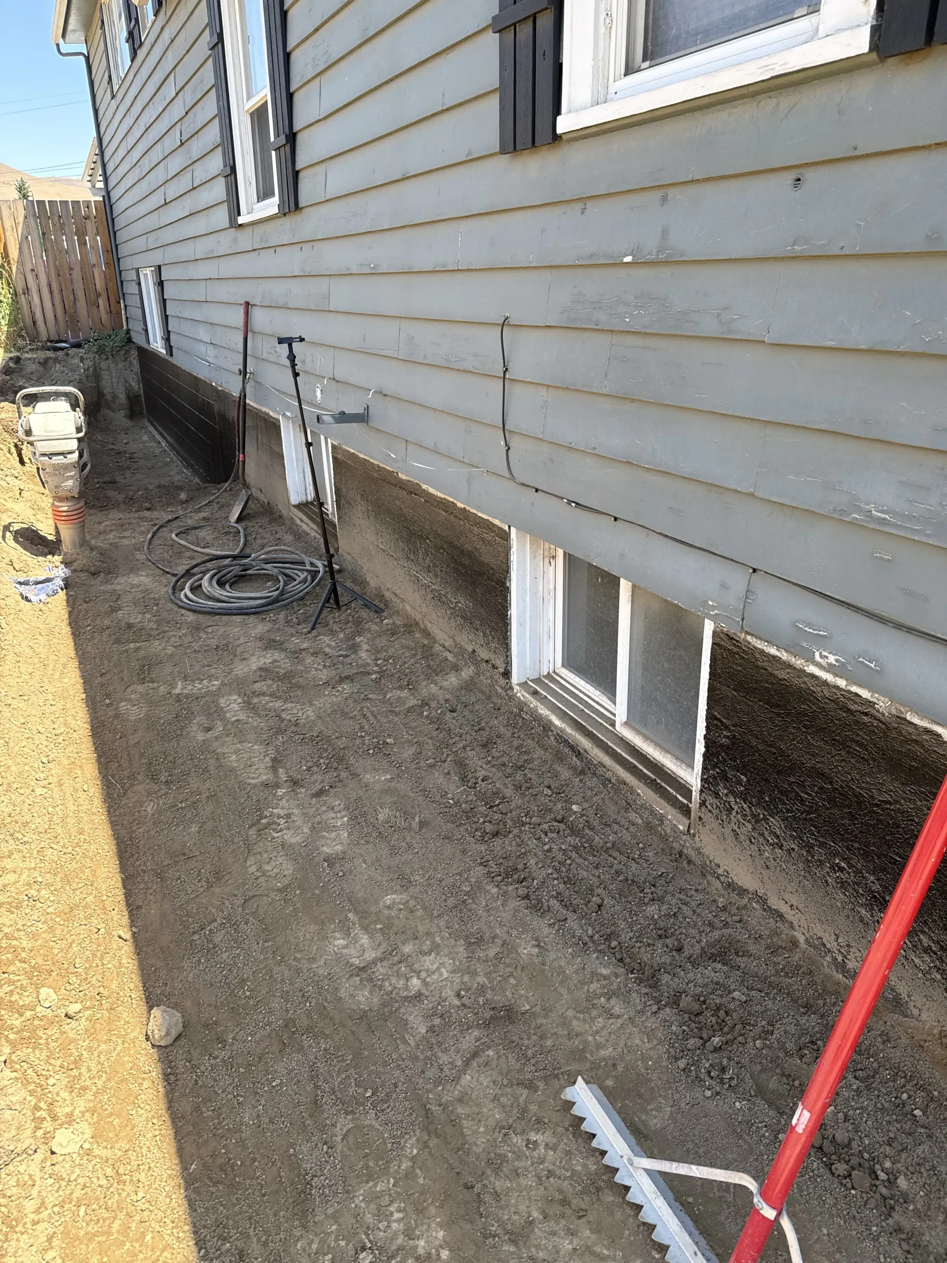 A ground-level view of a house foundation with grey siding, showing a dug-out trench, landscaping tools, and a window.