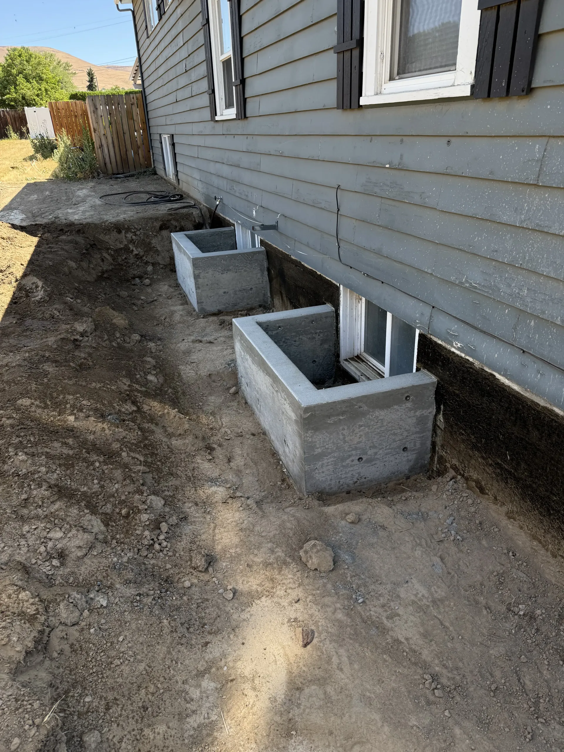 Two rectangular concrete window wells installed against the gray siding of a residential house with exposed dirt around.
