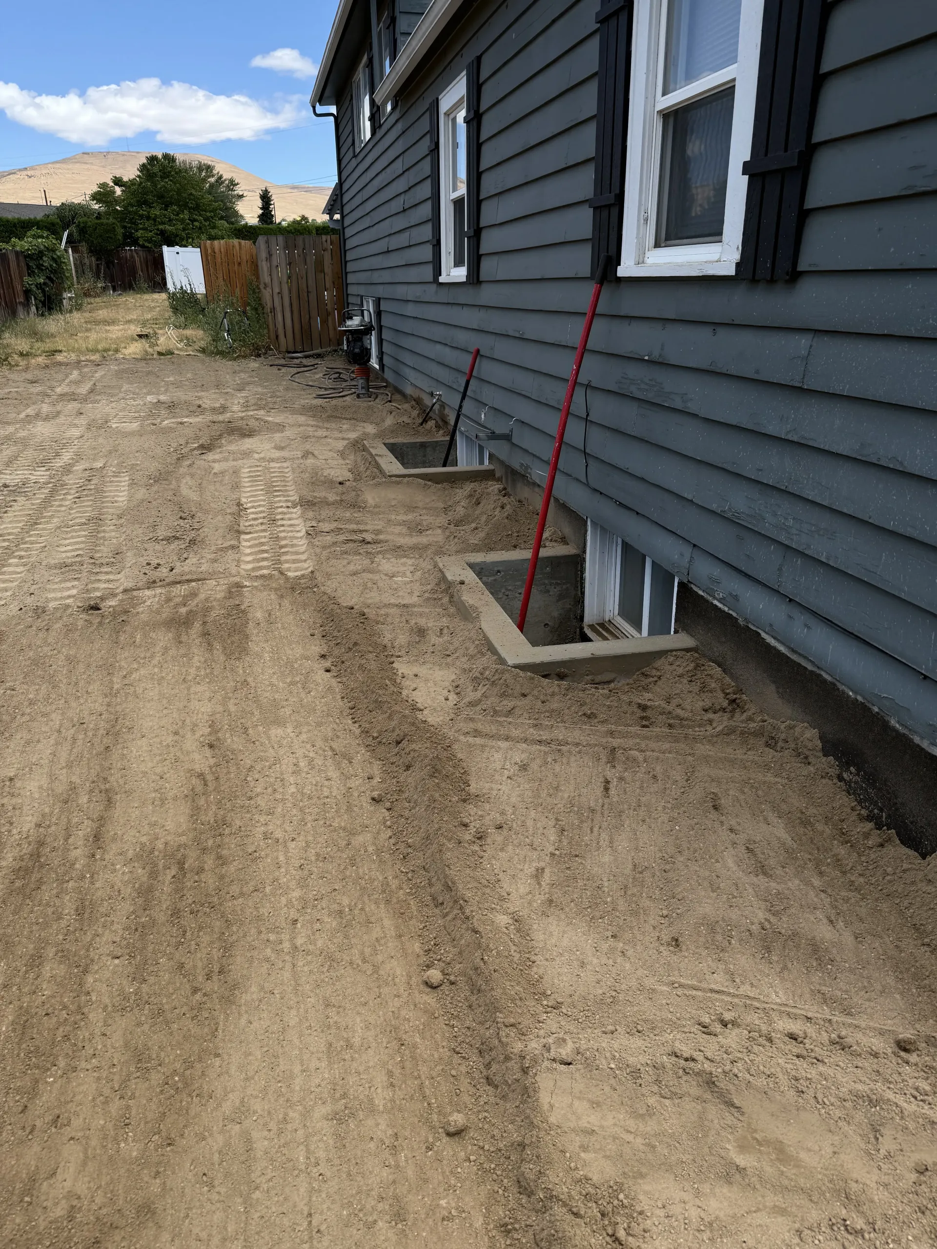 A side view of a gray-sided house with open foundation crawl space vents and a red tool resting against the wall.