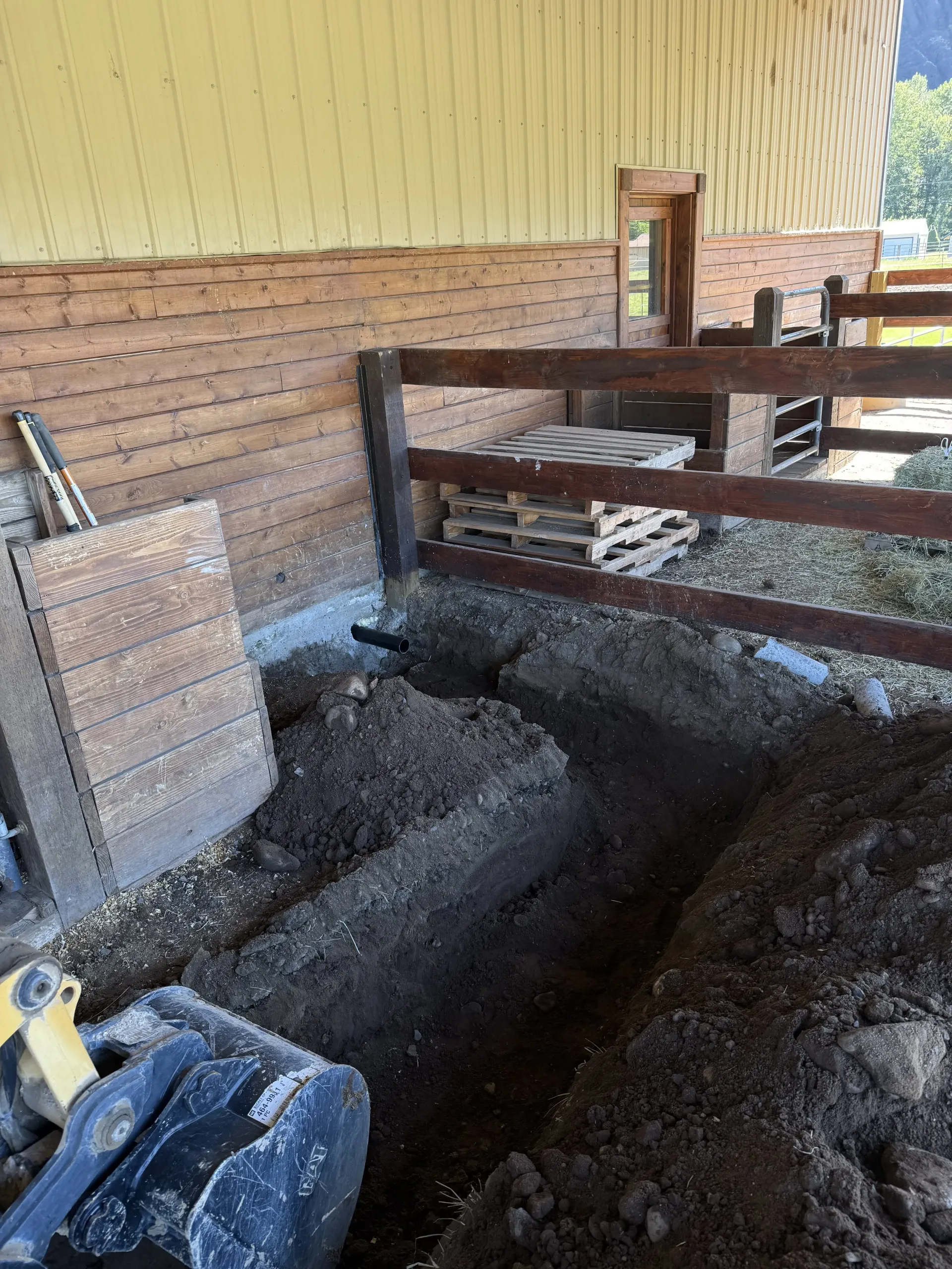A trench dug along the base of a tan building wall, next to a wooden fence and an excavator bucket.