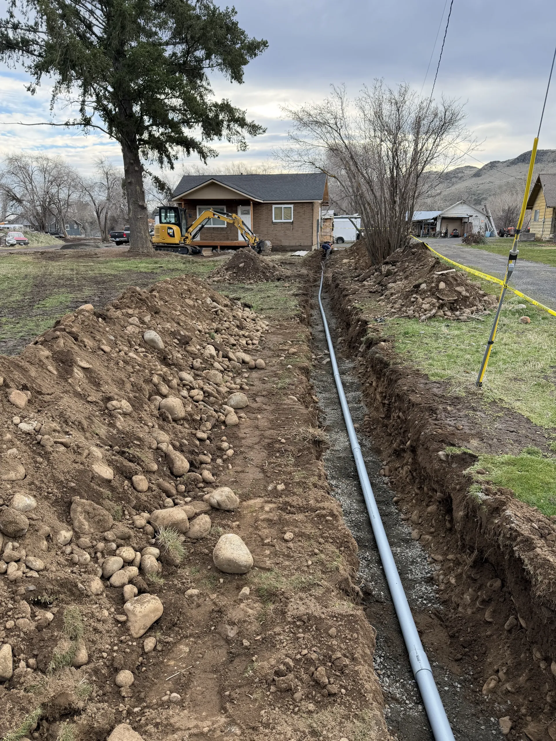 A trench with a gray pipe runs through a grassy yard toward a house under construction, with a small yellow excavator nearby.