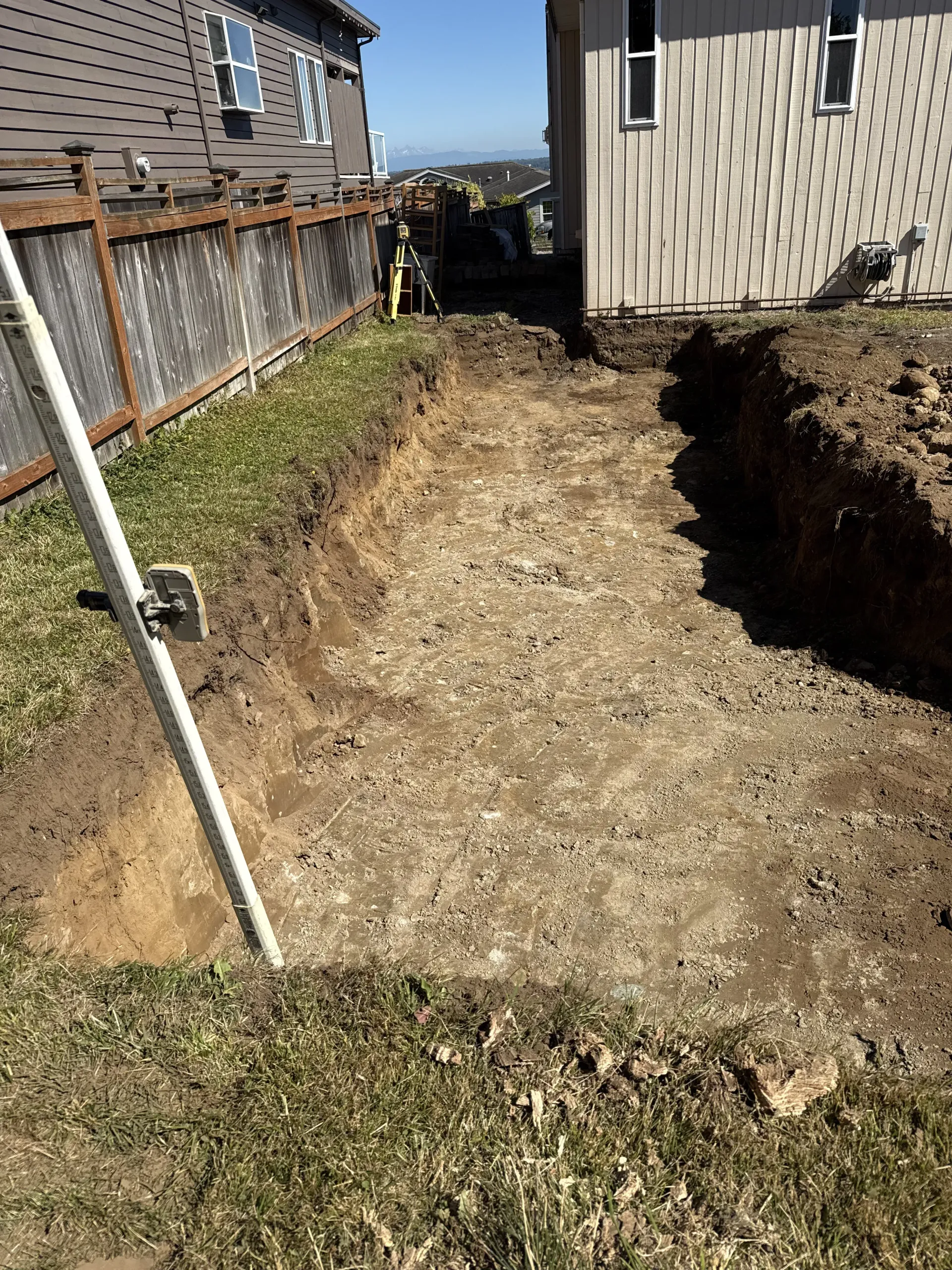 An excavated trench in a residential backyard between a house and a wooden fence on a sunny day.