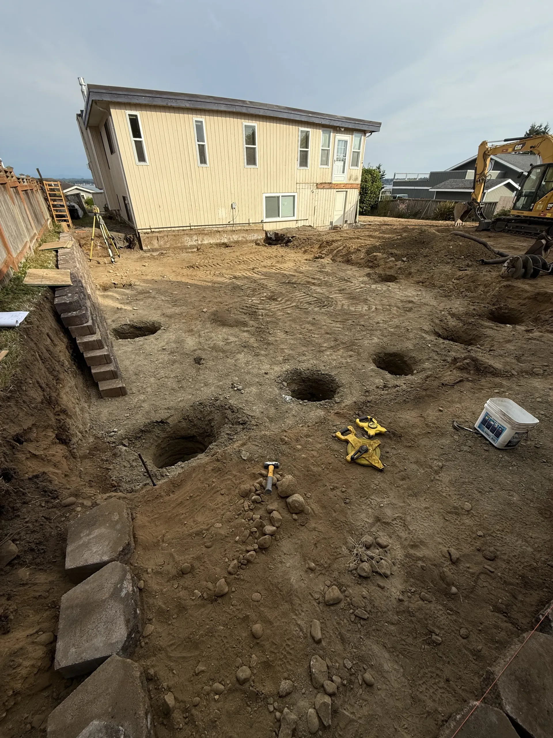 A residential construction site showing a house and open holes in the dirt ground for foundation piers.