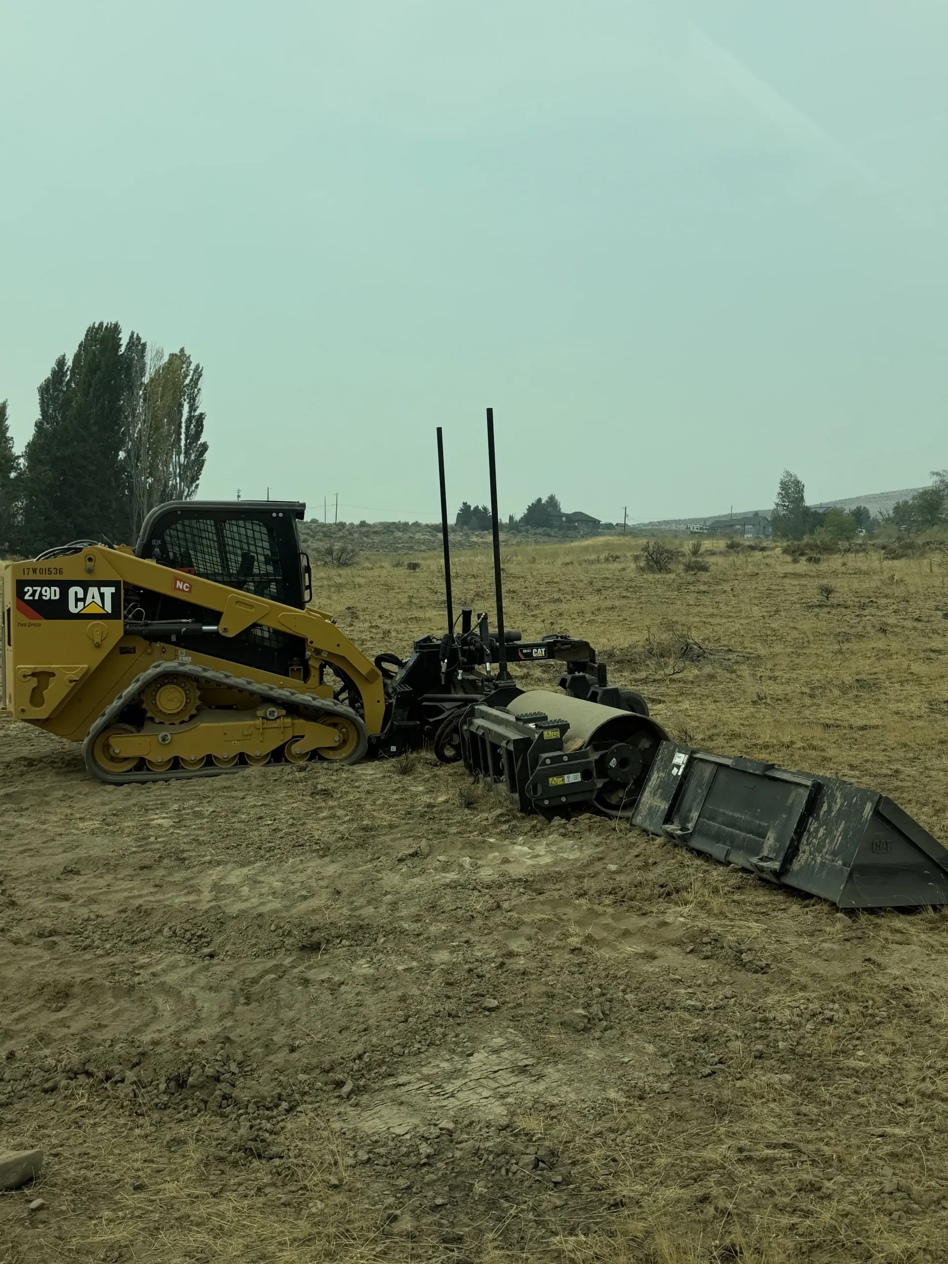 Yellow CAT compact track loader equipped with a forestry mulcher attachment working in a large, dry field.