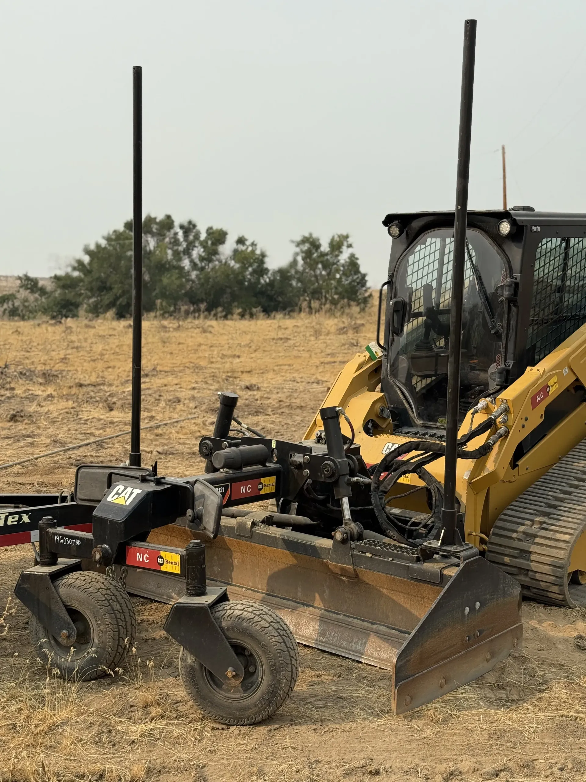 A yellow Caterpillar skid steer loader with a grader blade attachment parked in a field on a hazy day.