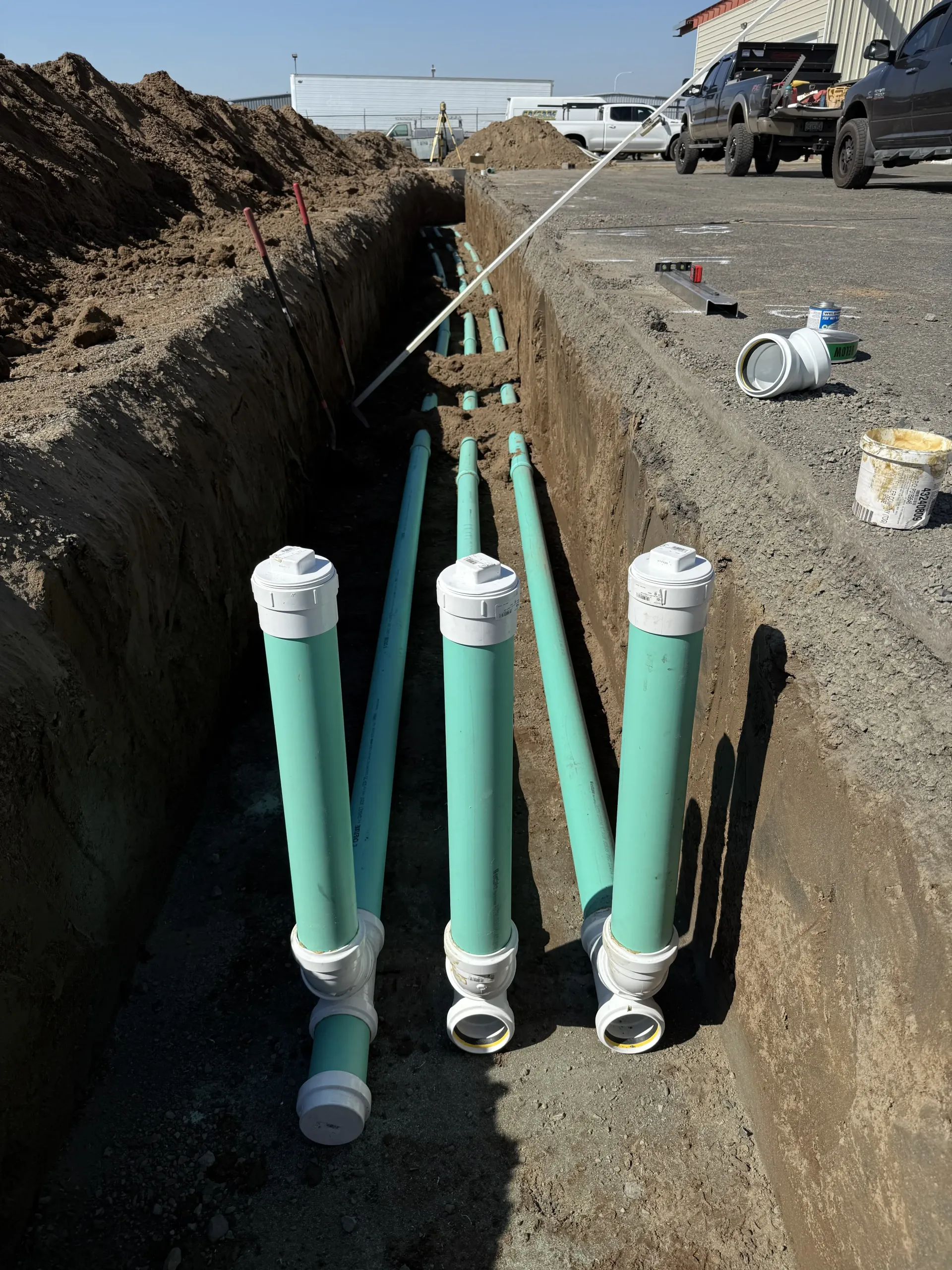 Three green PVC pipes with vertical extensions and capped ends installed in a long, narrow dirt trench at a construction site.