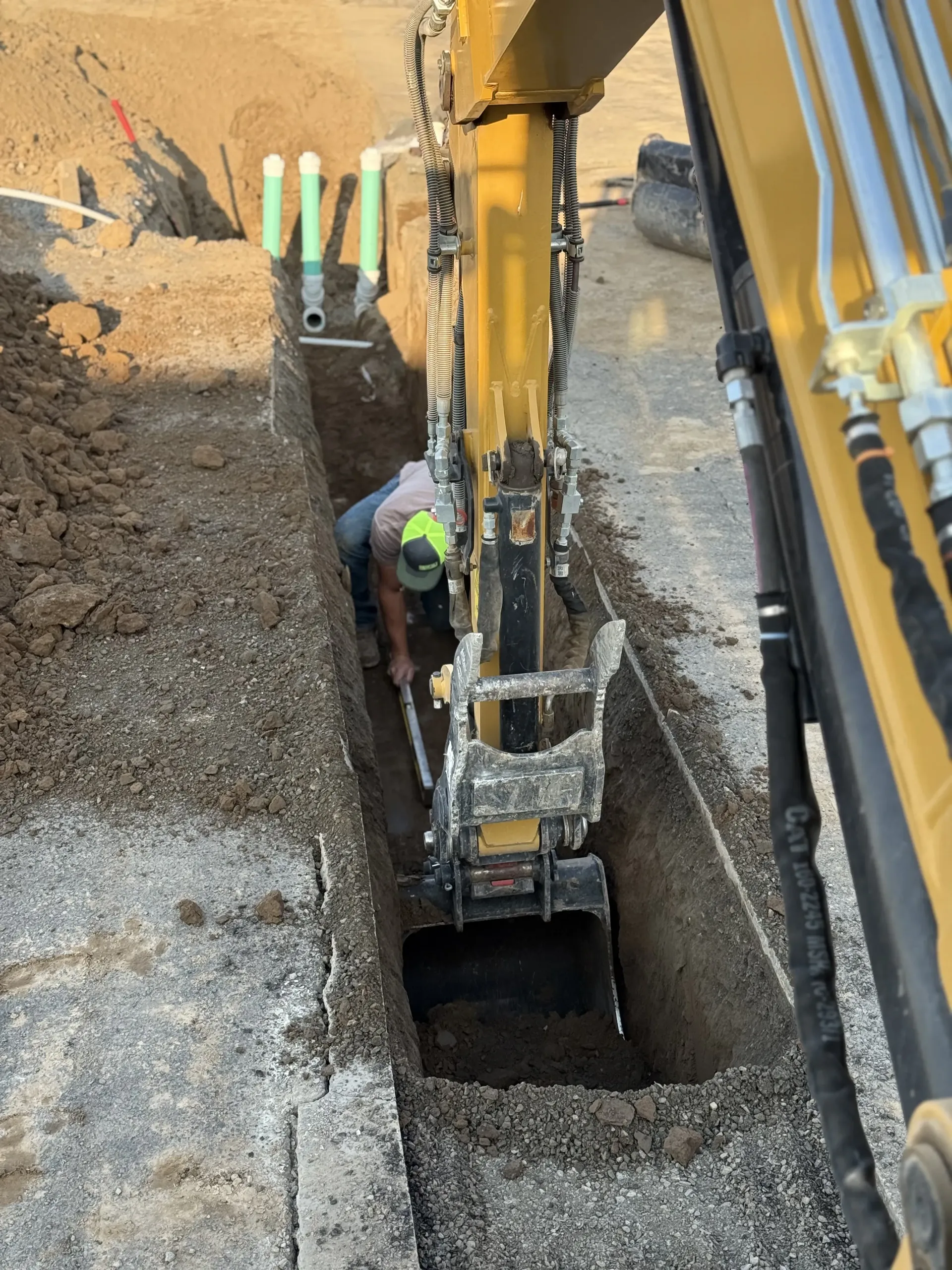 A person wearing a high-visibility vest works in a trench beside a yellow excavator's boom and bucket on a sunny site.