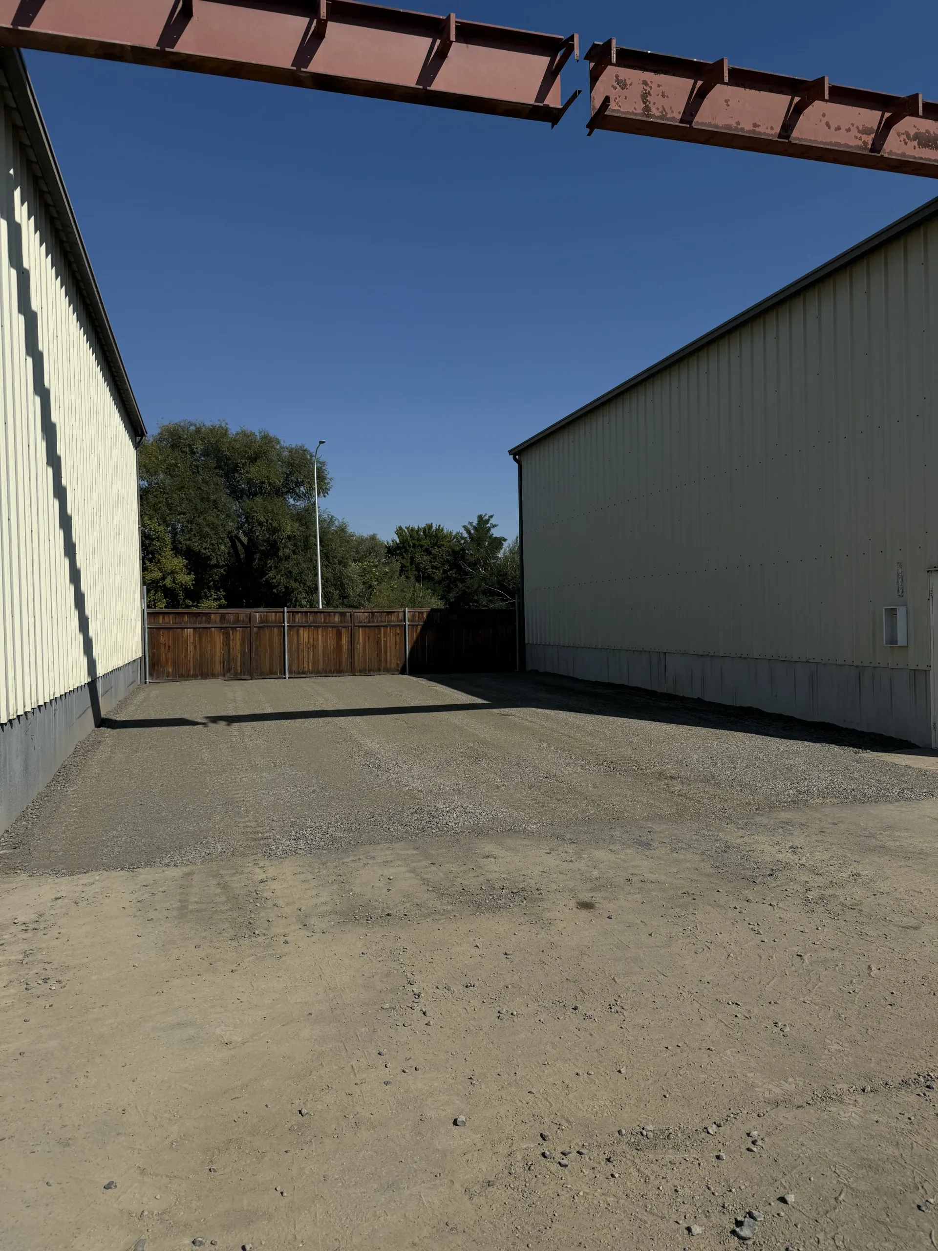 A gravel alleyway between two light-colored industrial buildings under a clear blue sky, with a wooden fence at the end.
