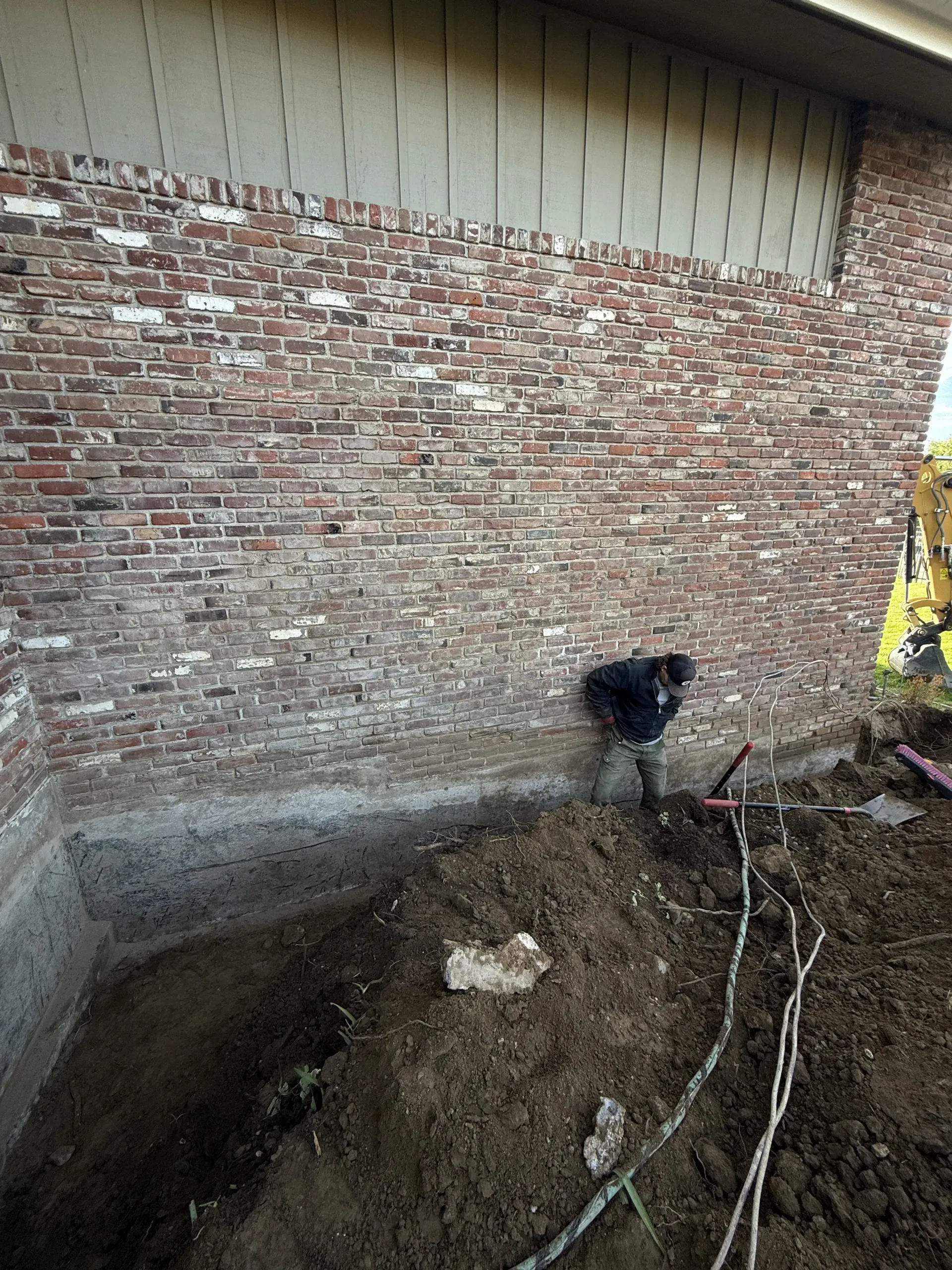 A worker stands in a deep trench against a brick building foundation during exterior construction work.