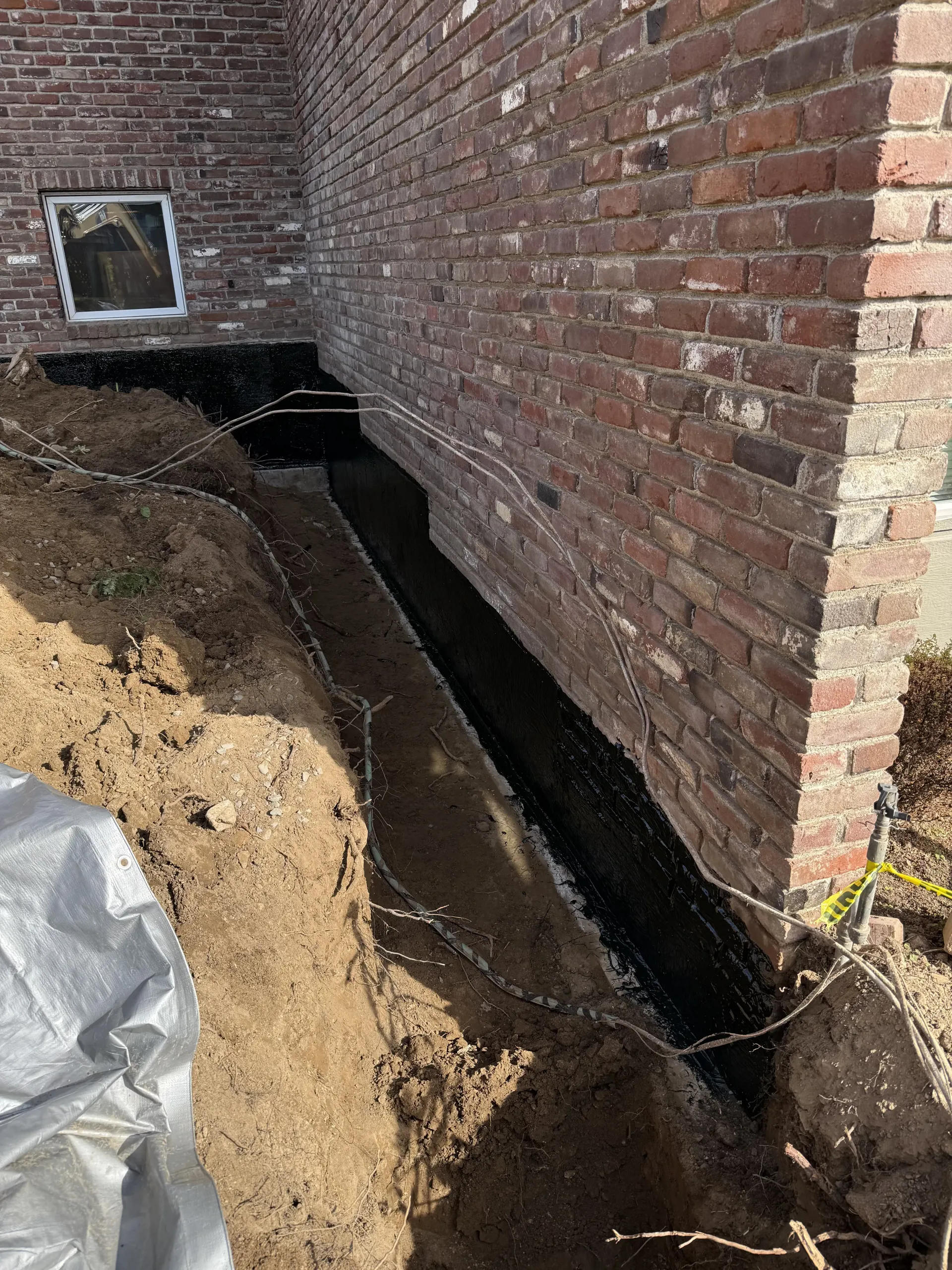 A brick house foundation exposed by a deep trench, coated with black waterproof sealant.