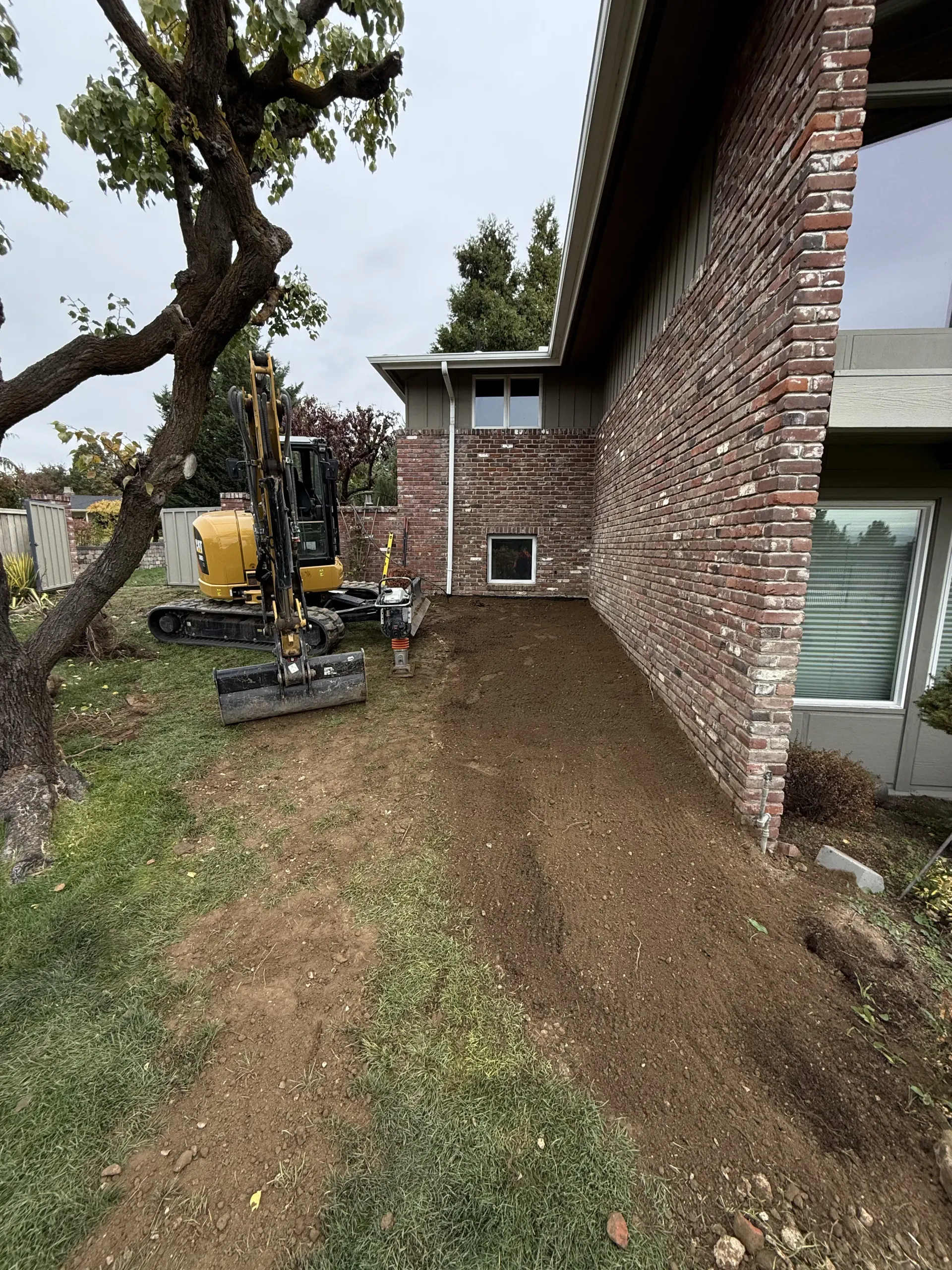 A yellow excavator parked on soil next to the side of a brown brick house during a landscape construction project.