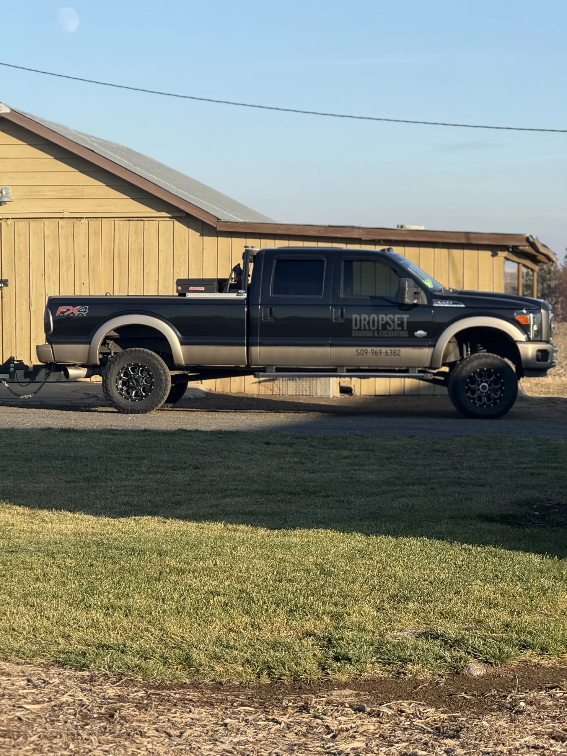 A dark-colored, lifted pickup truck with gold-tone lower trim parked on gravel in front of a tan barn.