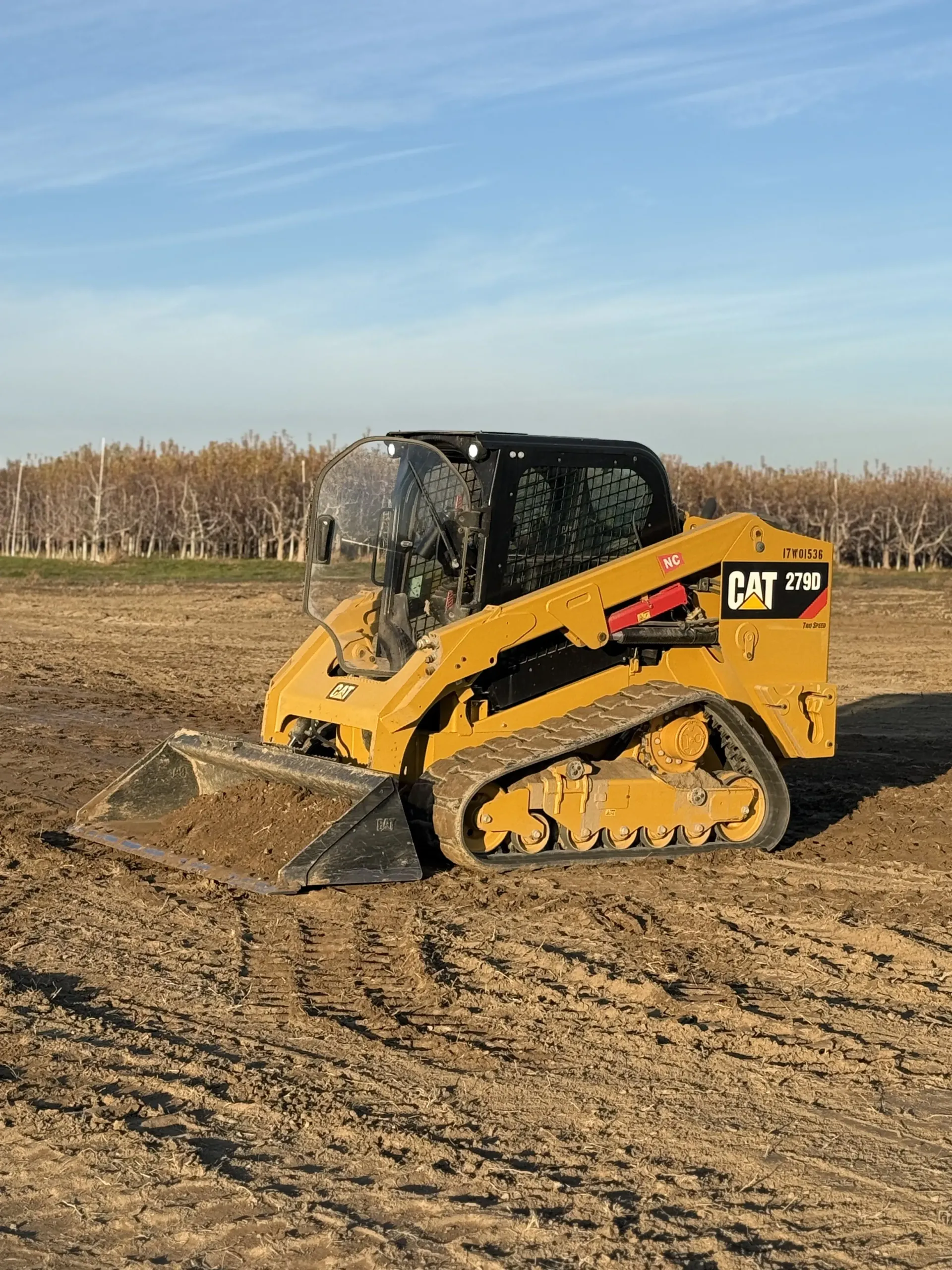A yellow Caterpillar compact track loader sits in an open, dirt-covered field under a clear blue sky.