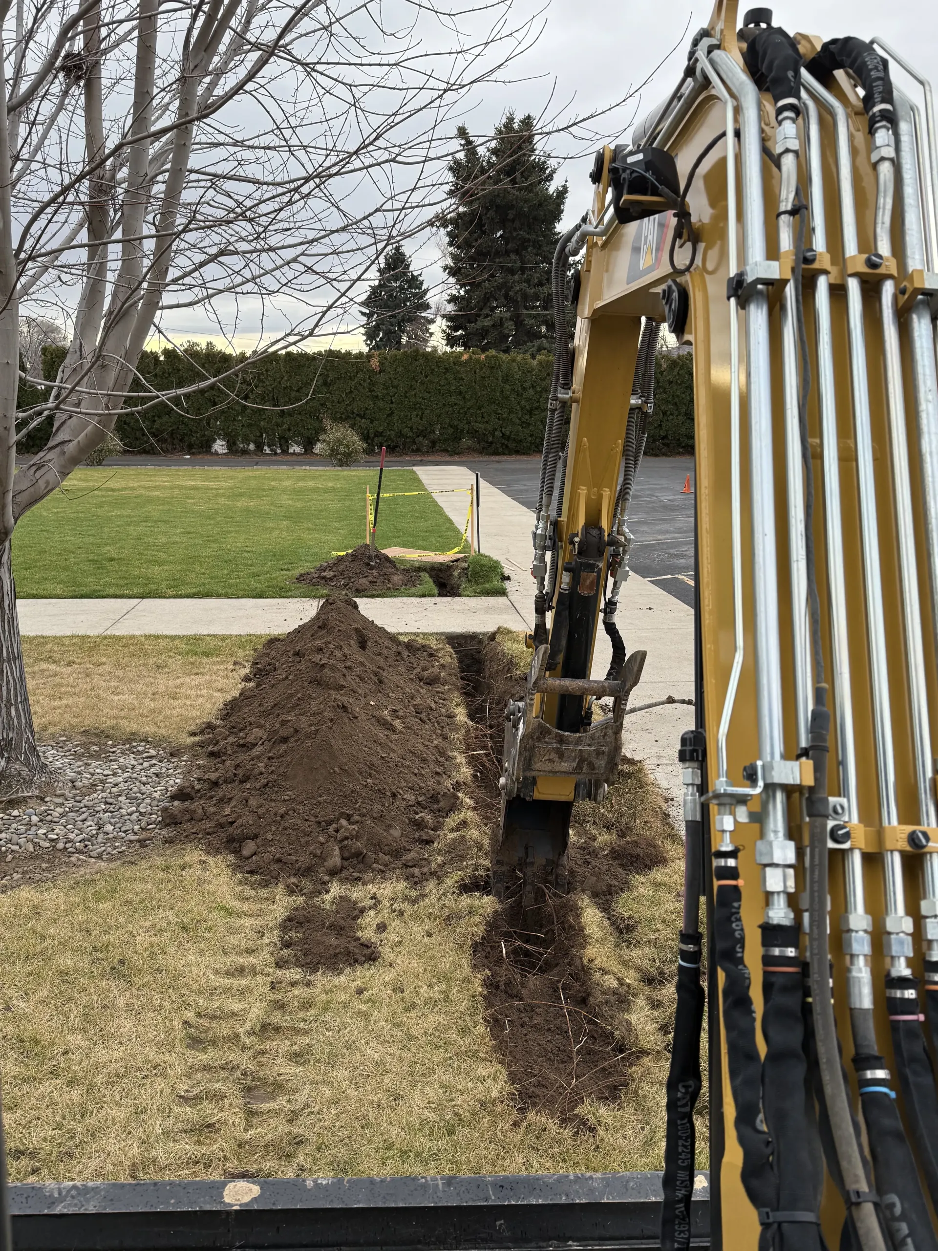A yellow construction excavator digs a trench in a grassy yard, with a pile of dirt beside the trench.