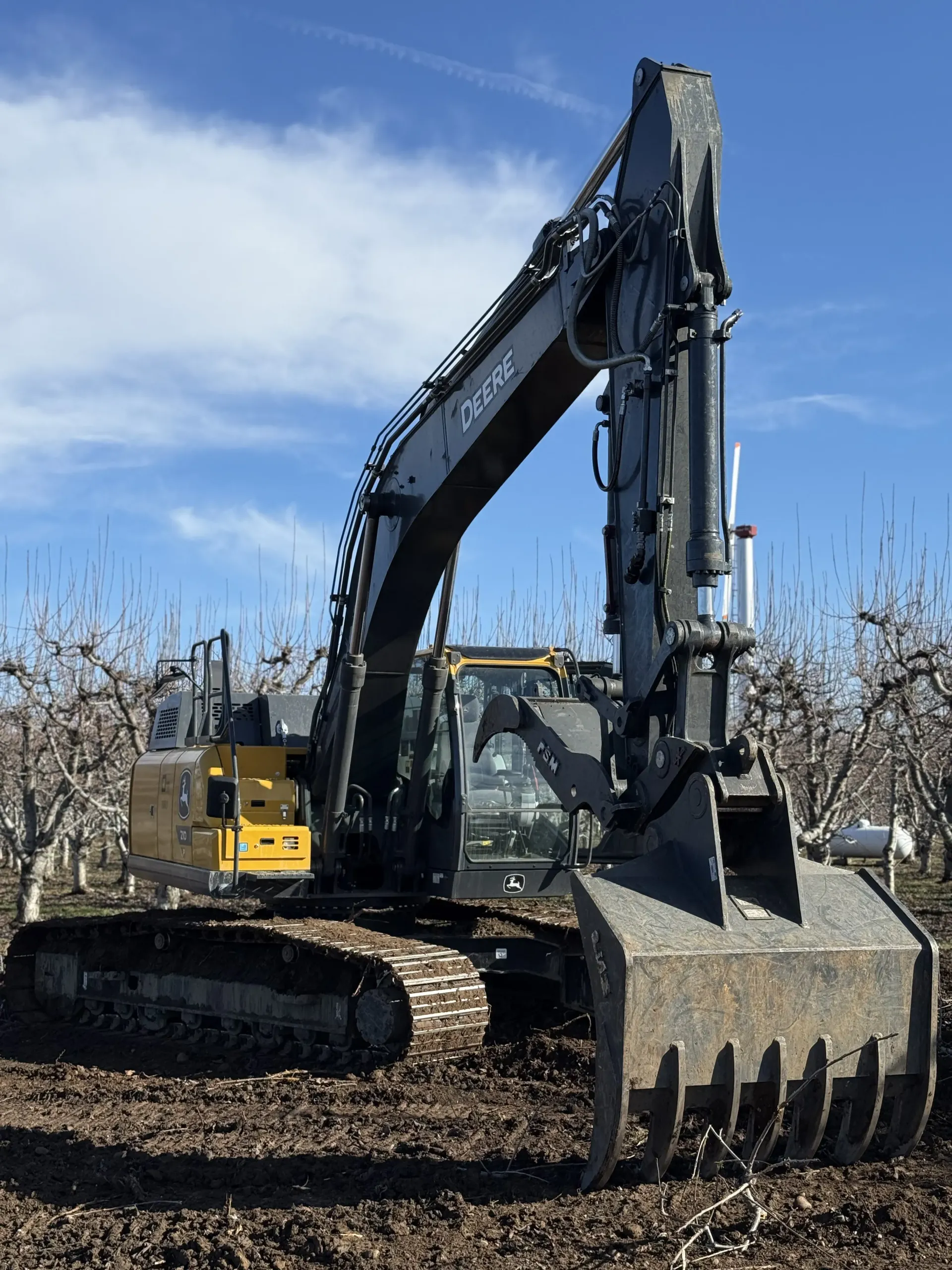 A yellow and dark gray John Deere track excavator equipped with a large grapple in an orchard.
