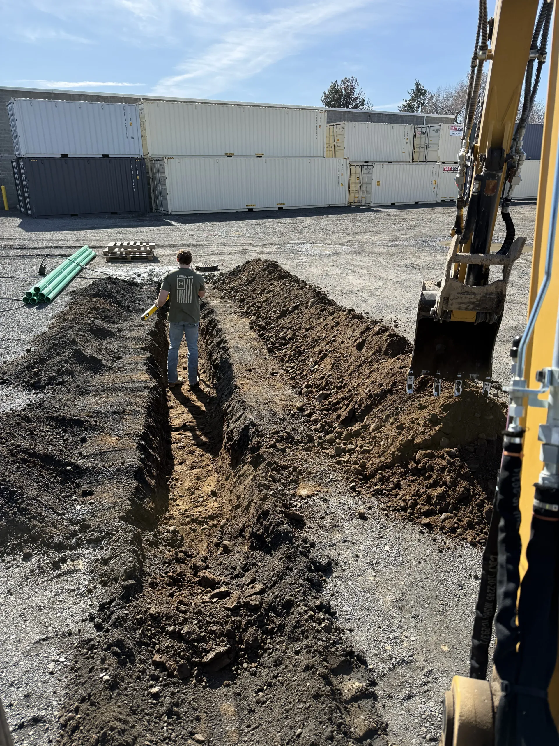 A construction worker stands by a trench in a rocky yard, with an excavator arm visible to the right.