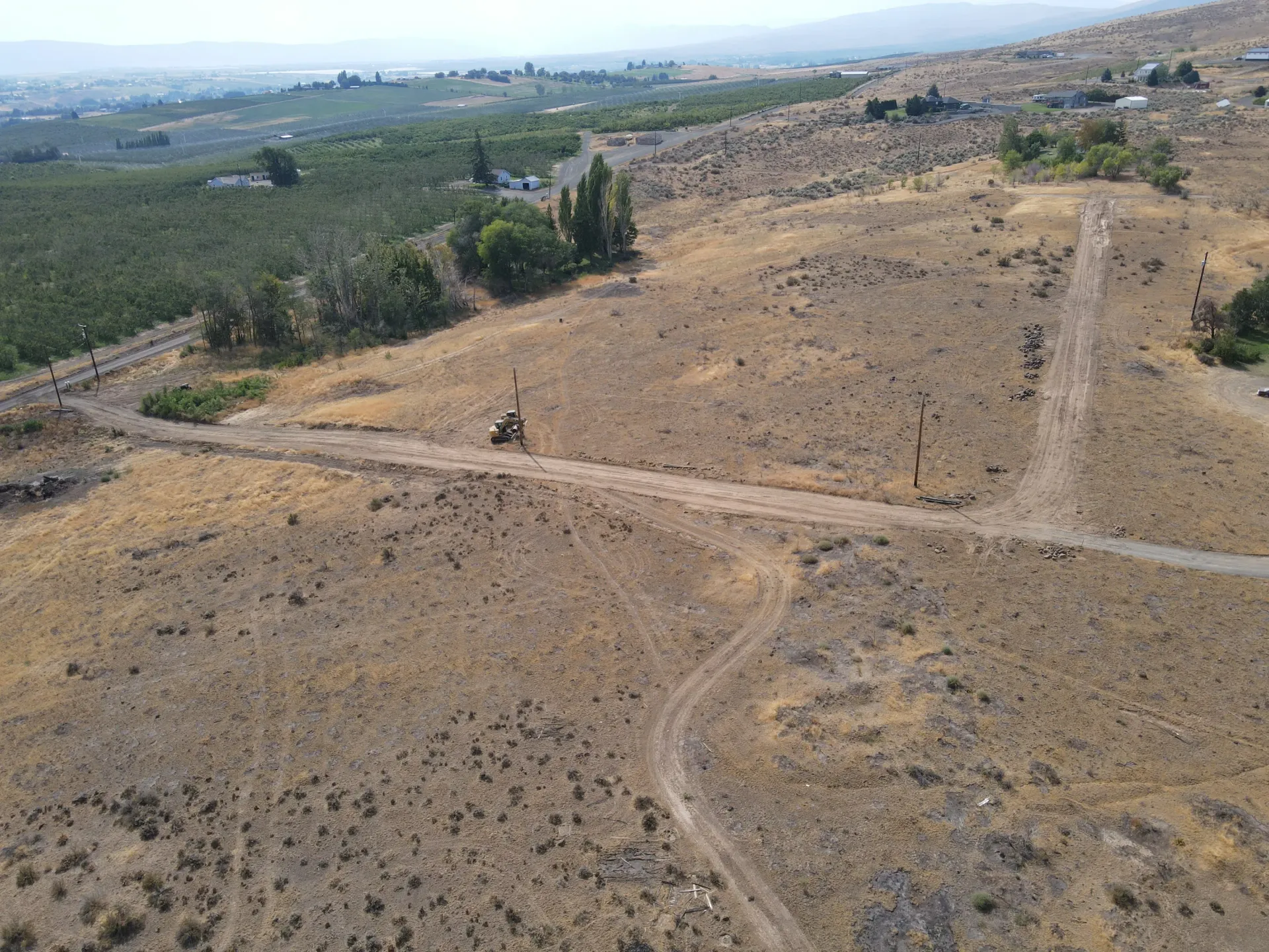 An aerial view of a dry, arid hillside with dirt paths traversing the landscape next to a lush, green orchard.