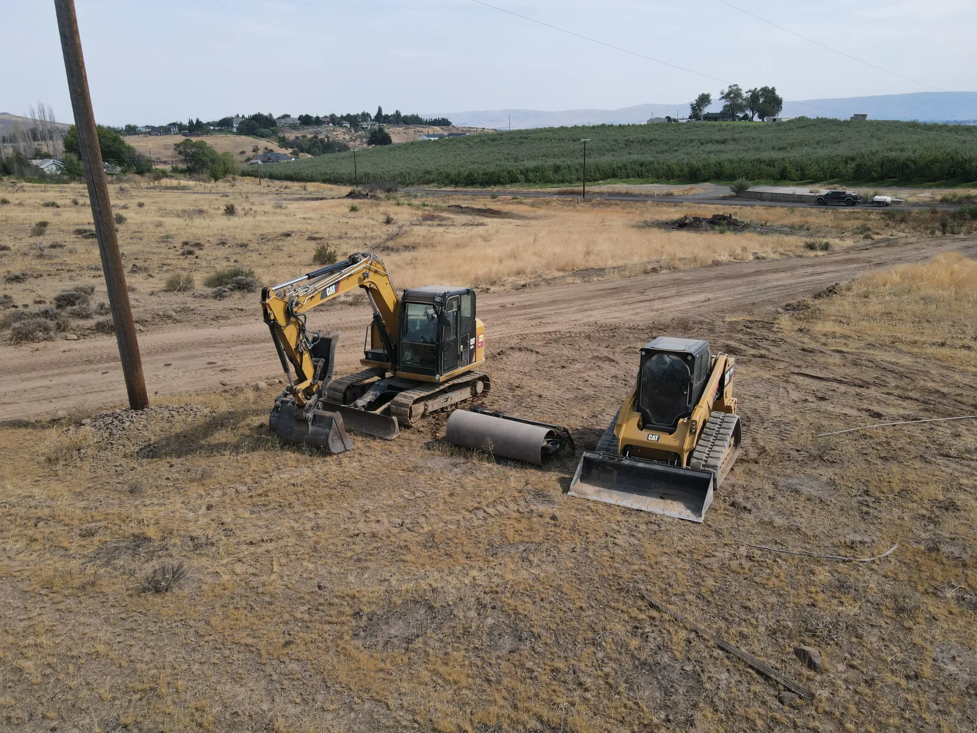 A yellow excavator and a skid steer parked on a dry, dirt lot next to a utility pole, with green fields in the distance.