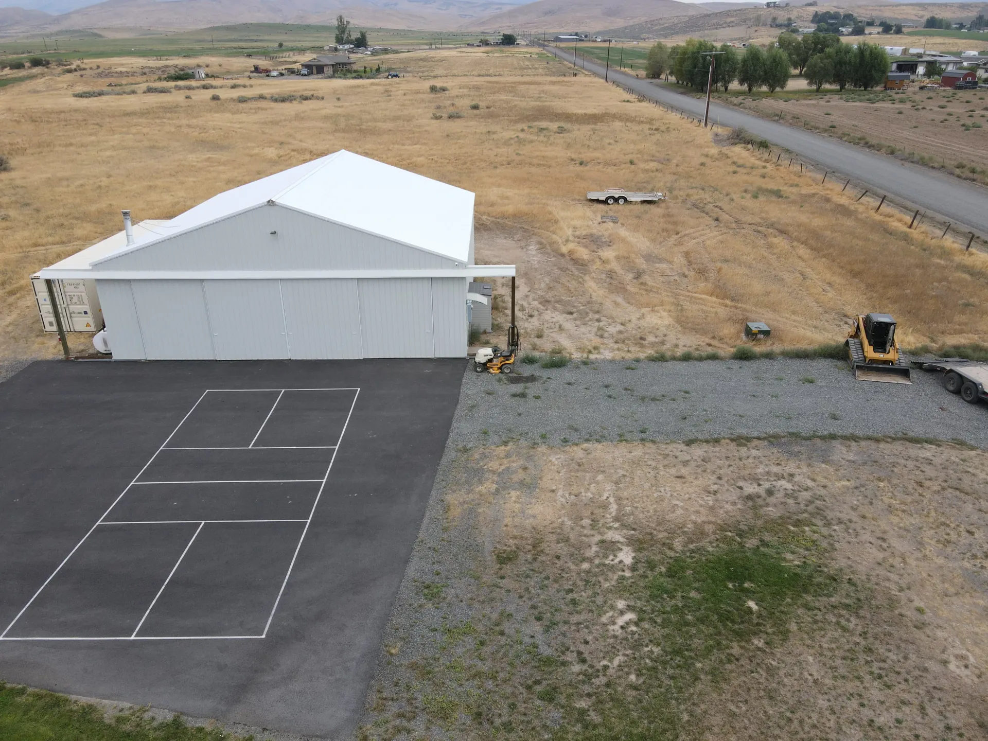 An aerial view of a white hangar building with a black asphalt driveway and pickleball court in a rural field.
