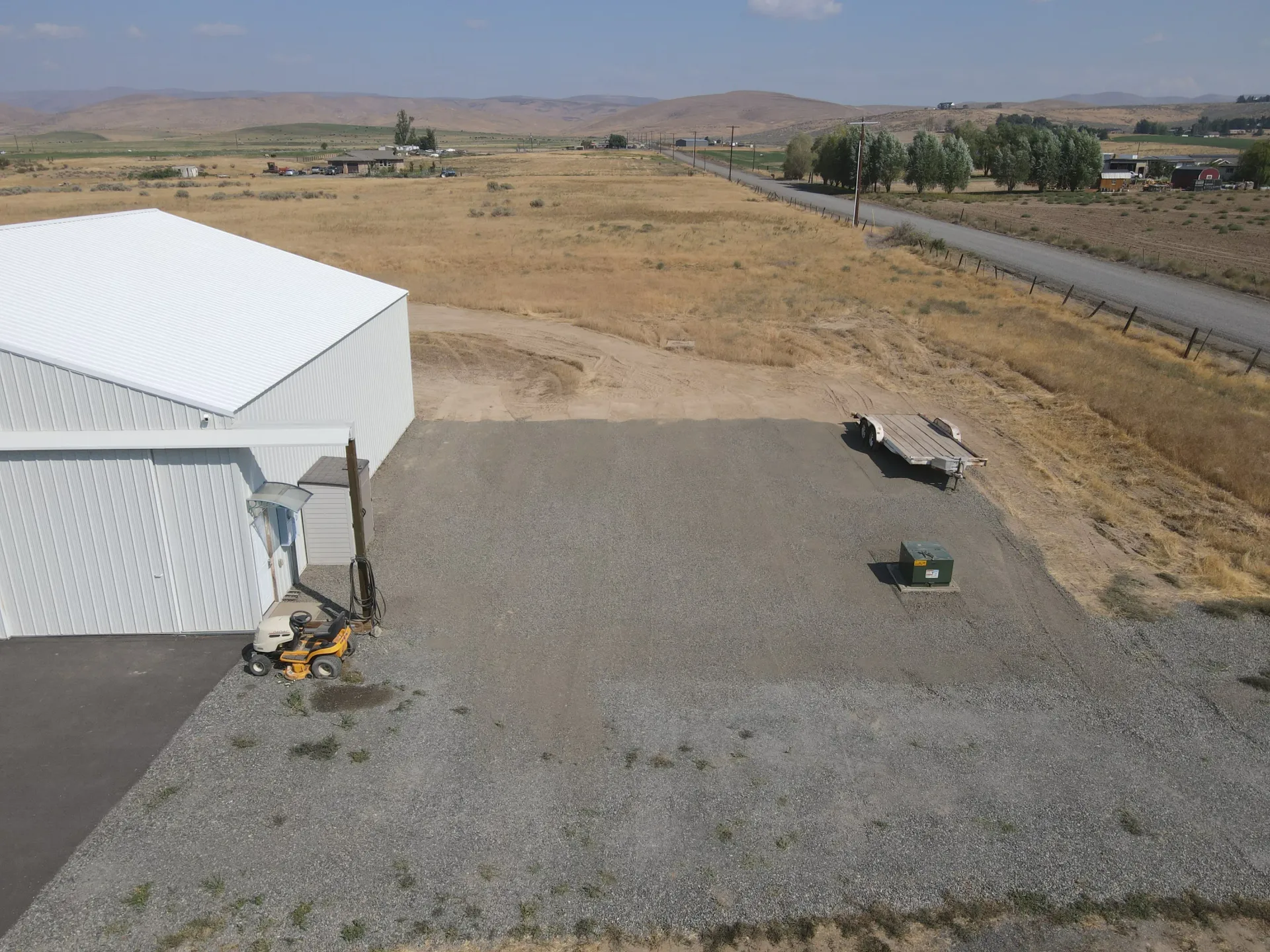 A white building stands next to a gravel yard, with a yellow piece of construction equipment and a road in the distance.