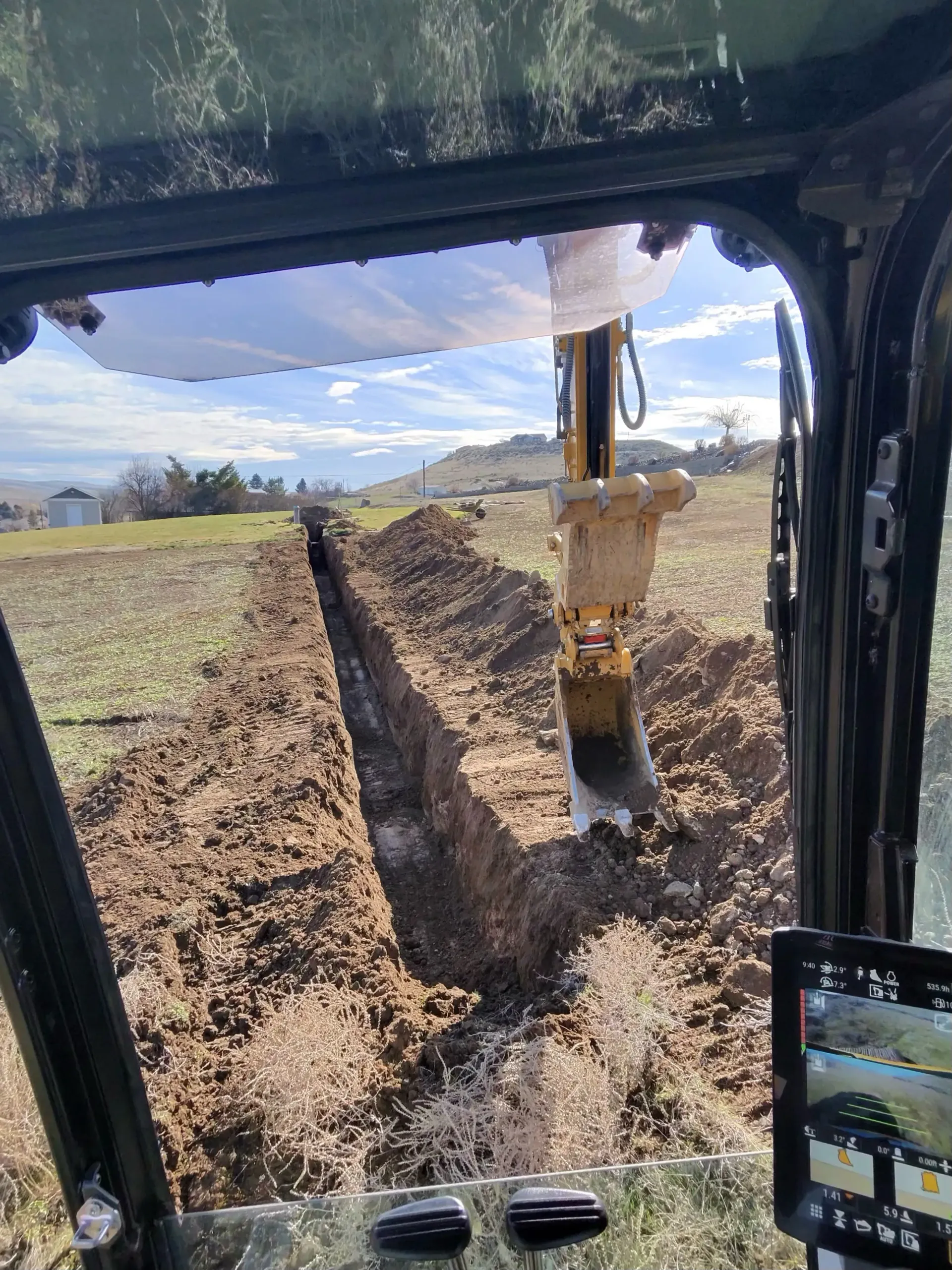 View from inside an excavator cab looking out at a deep trench dug into an open, sunny field.