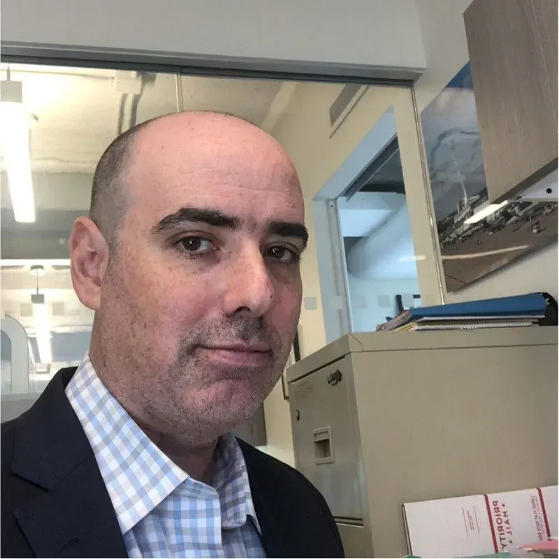 Man standing in an office beside filing cabinets, wearing a dark jacket and checkered shirt.