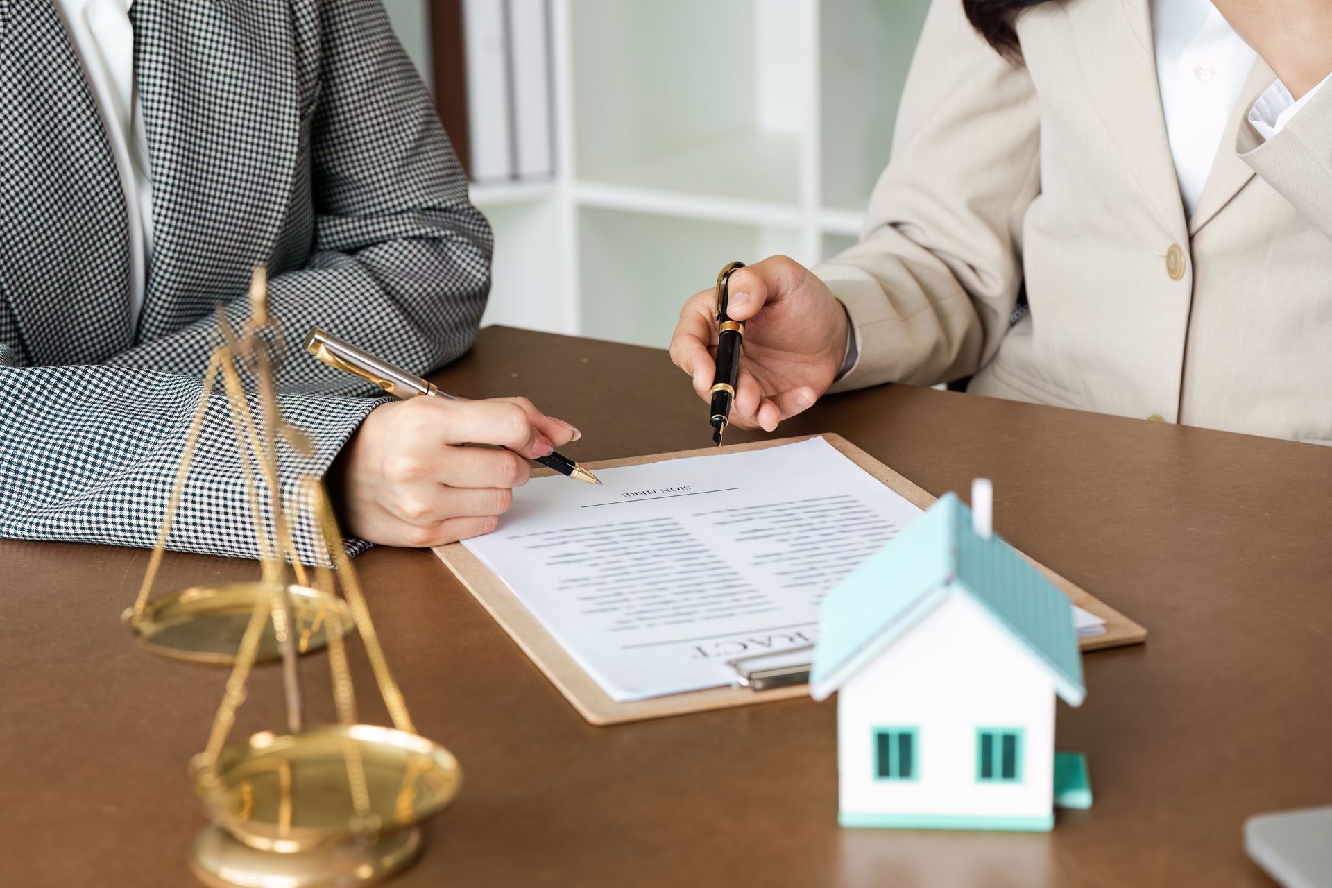 Two people signing a document at a table with a small house model and scales of justice