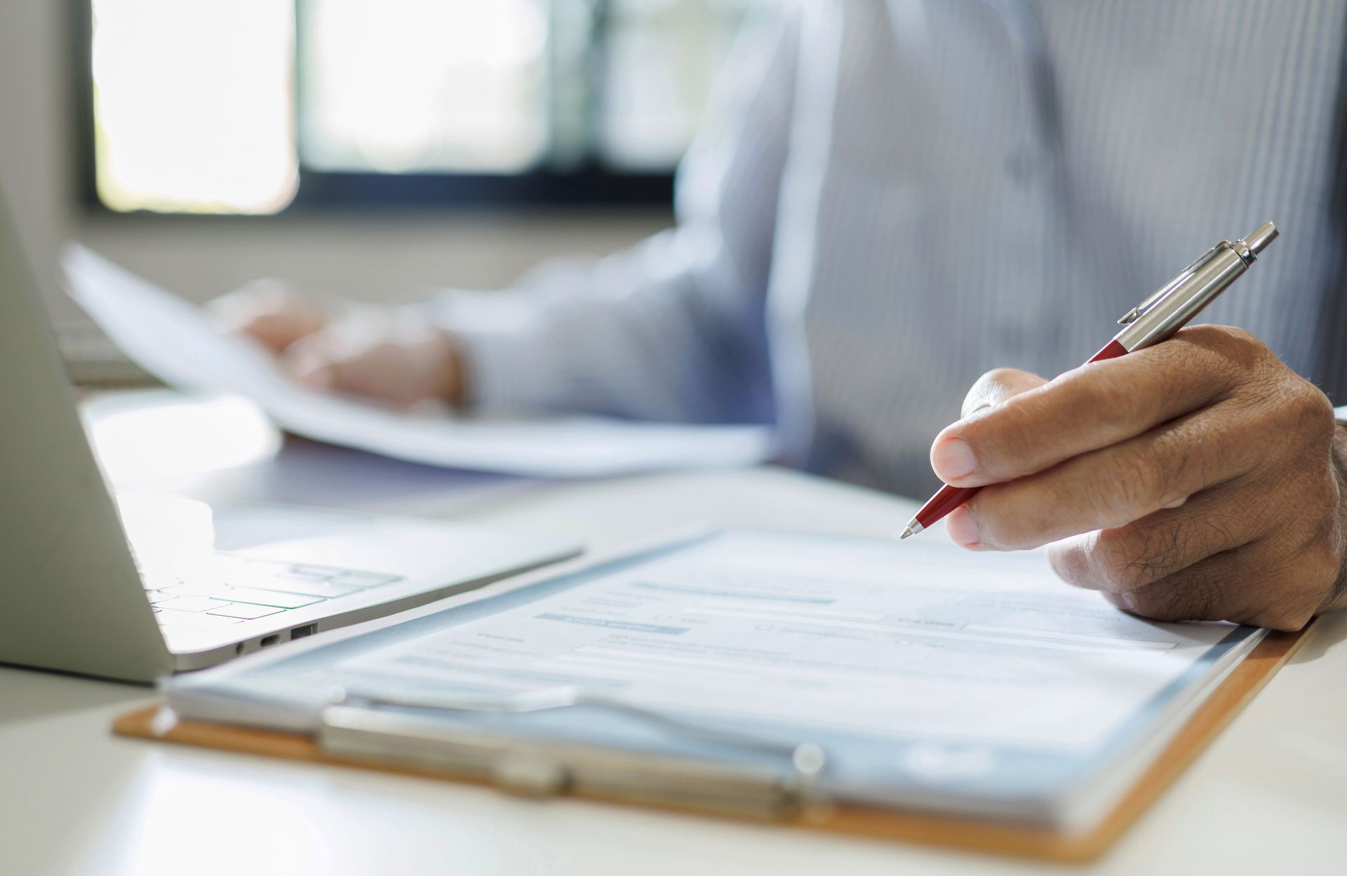 Hand writing on a clipboard beside an open laptop in an office setting