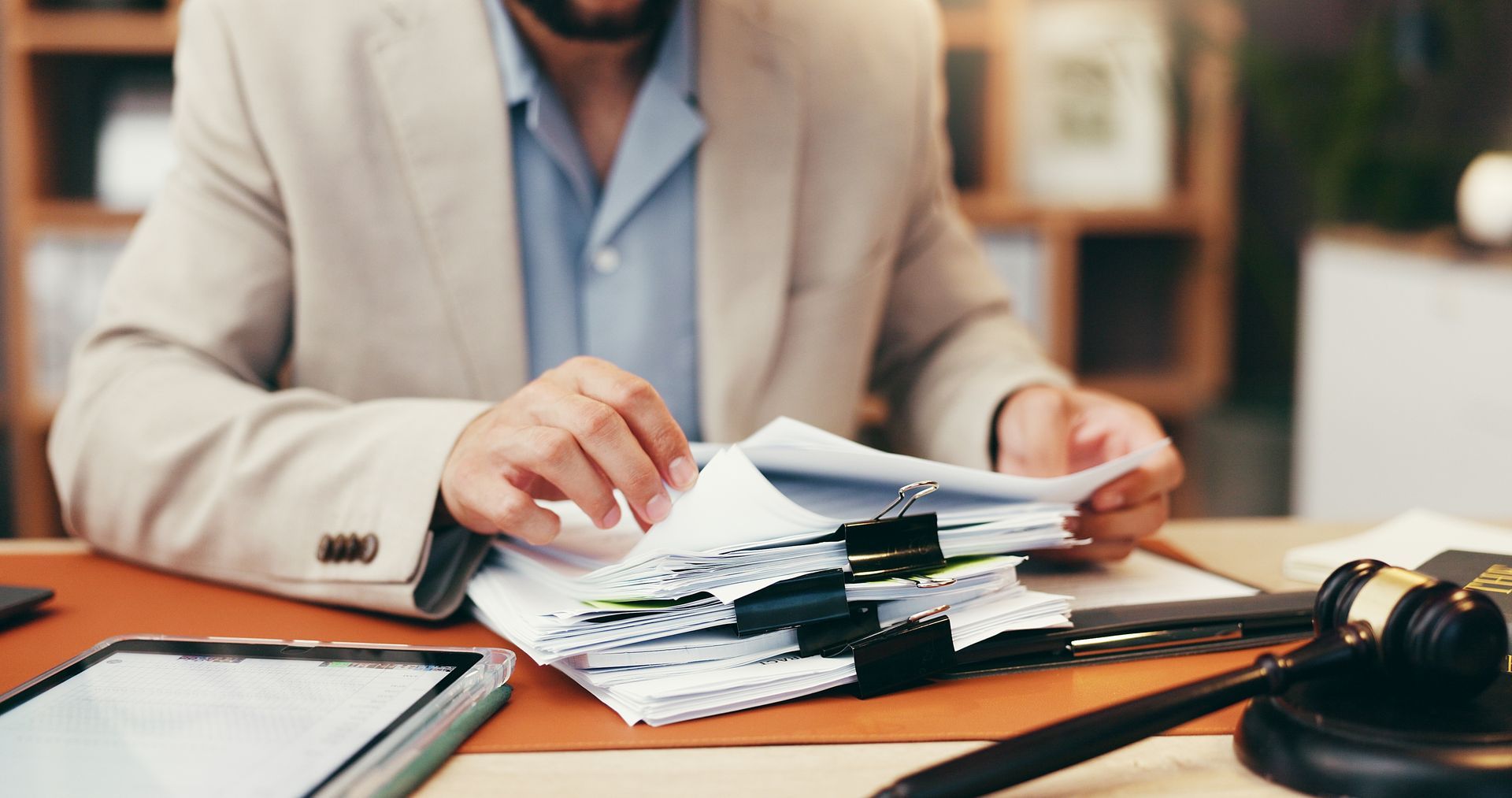 Person in a beige blazer reviews a stack of documents at a desk with a laptop and gavel in view