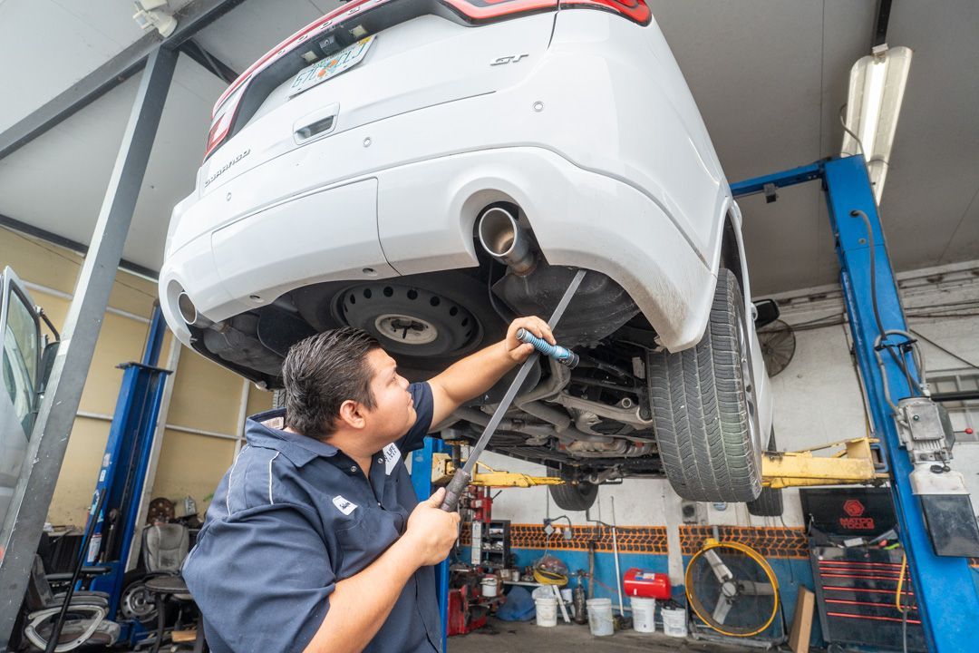 Mechanic working on the underside of a white SUV lifted on a blue hydraulic lift in a garage.